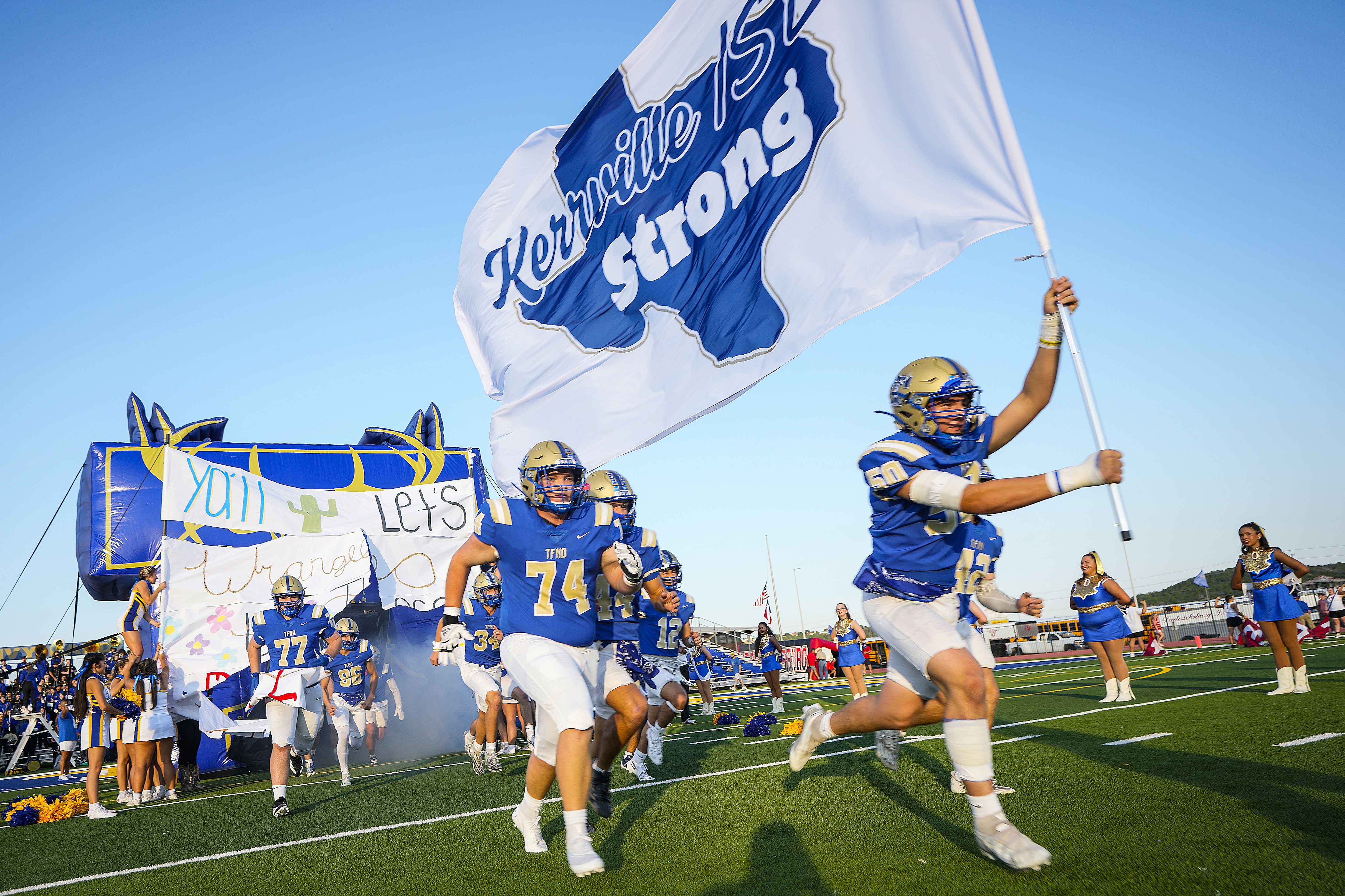 Tivy High School football players run onto the field before their game against Fredericksburg at Tivy Antlers Stadium in Kerrville in September 2025.  The lead player carries a sign that reads "Kerrville strong" over a blue-colored Texas. 