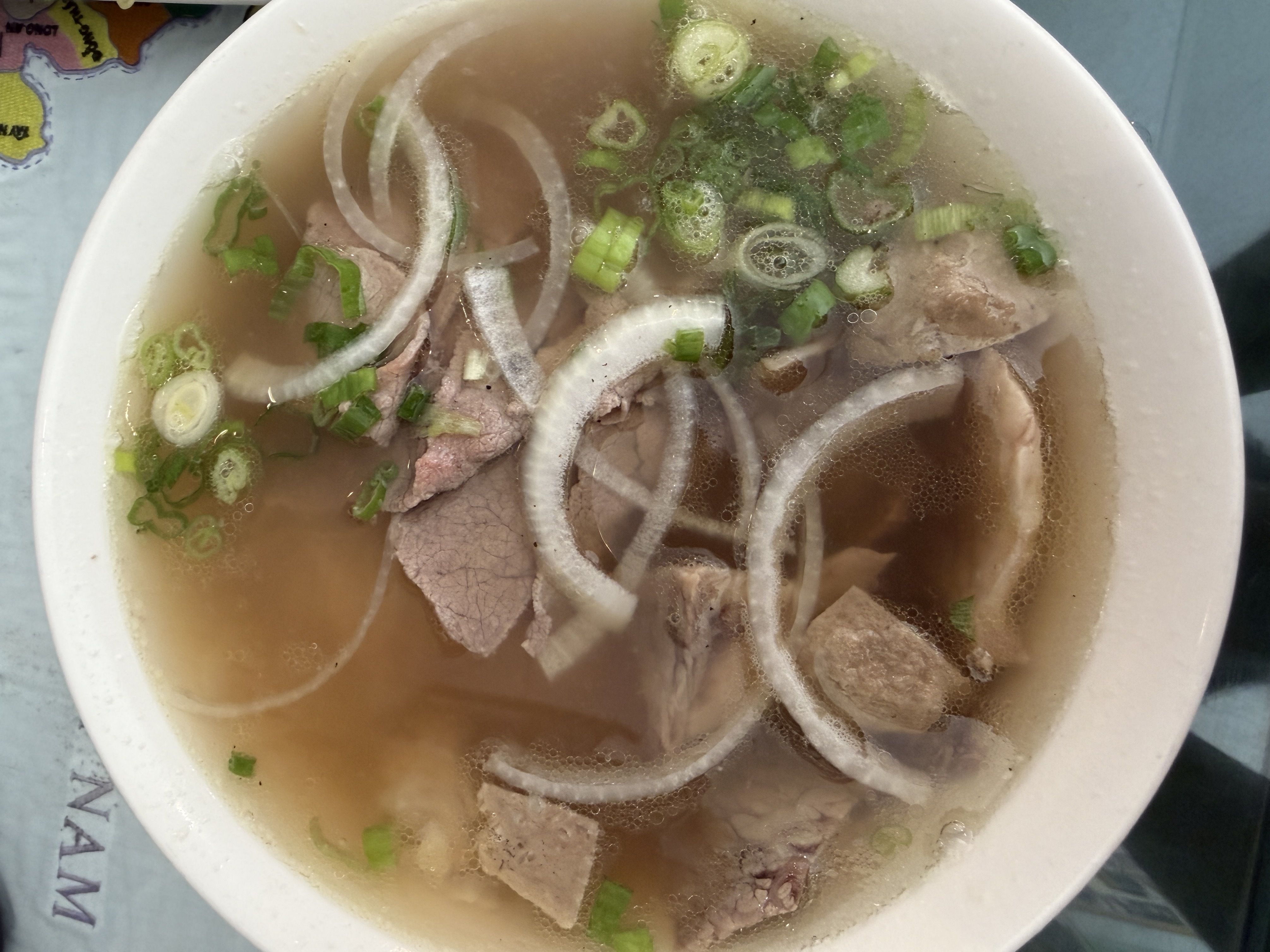 Top-down view of a white bowl of clear broth soup with green onion pieces, thin slices of pork, and onion rings floating in a light brown broth.