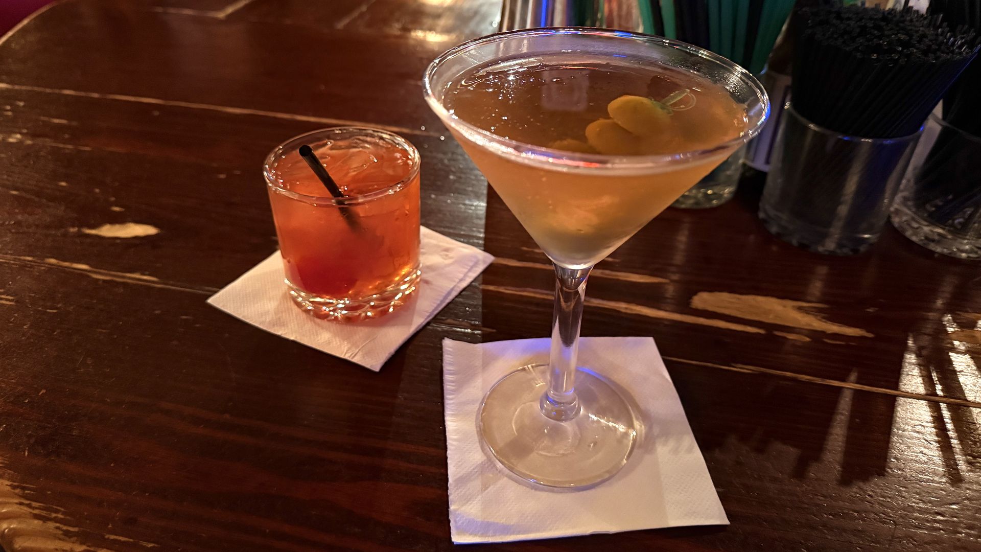Two cocktails on a dark wooden bar: a large martini glass and a short orange drink with a straw on a napkin, with green straws and bar bottles in the background.