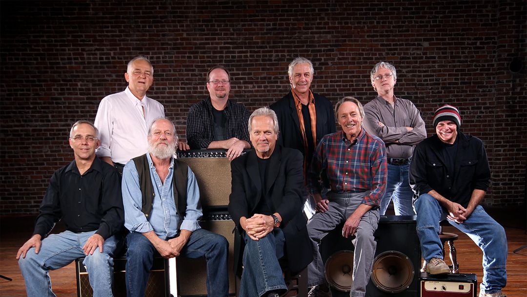 A music group of nine men sit around speakers and amps, posing for the camera. 