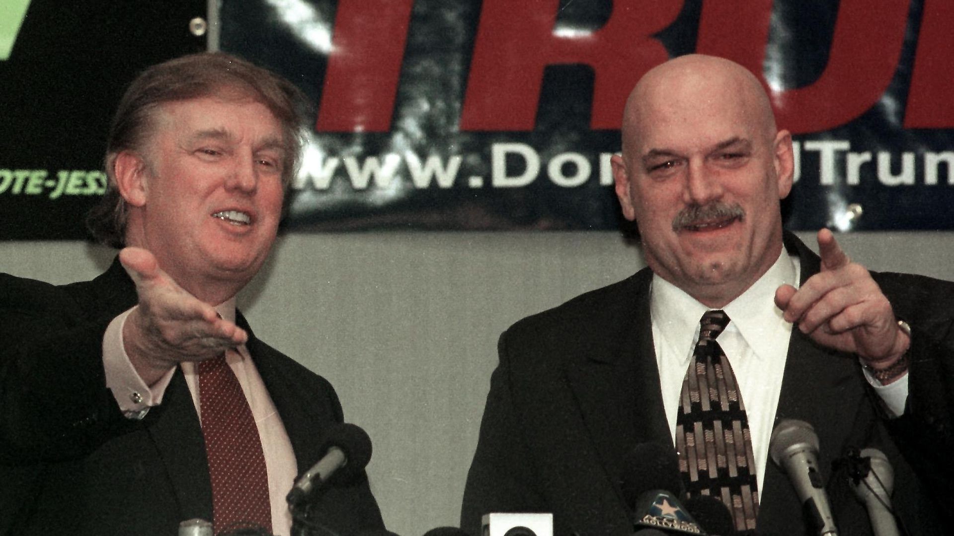 Two men stand behind a row of microphones and gesture toward the audience unseen in the foreground