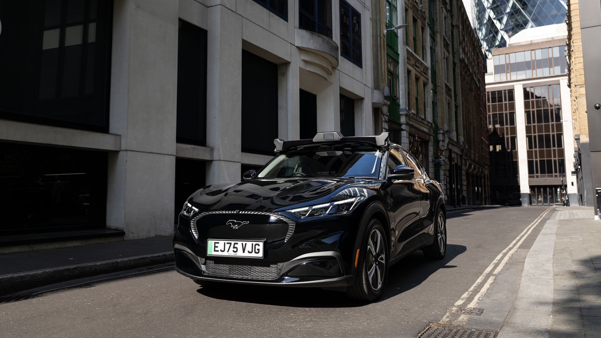 Black Mustang Mach-e with autonomous vehicle sensors on the roof, parked on a narrow city street, flanked by tall beige and brick buildings.