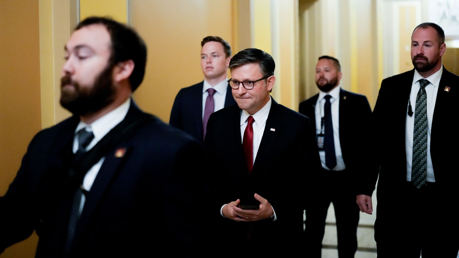 House Speaker Mike Johnson, wearing a dark gray suit and holding a phone while flanked by Capitol Police officers in a yellow hallway.