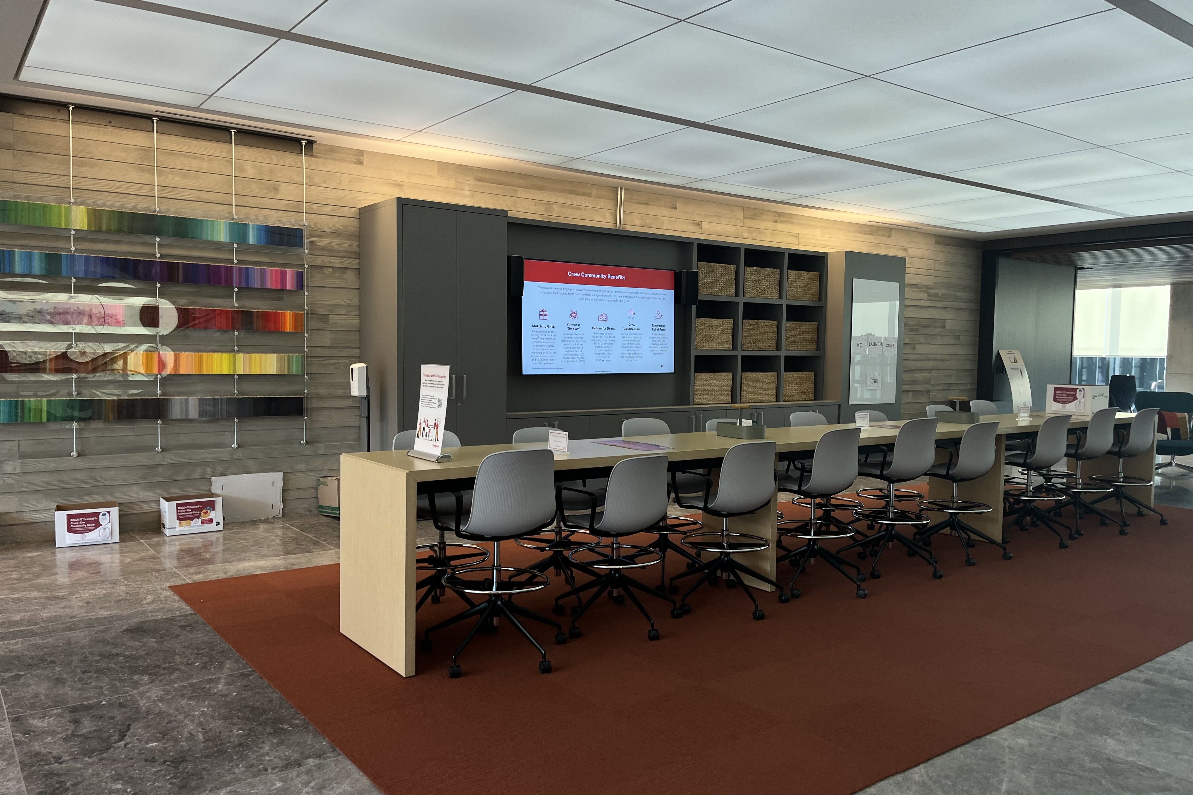 Modern room with a long light wood table and gray chairs on a rust-colored carpet. Gray cabinets with baskets and a digital display board on a wood panel wall.