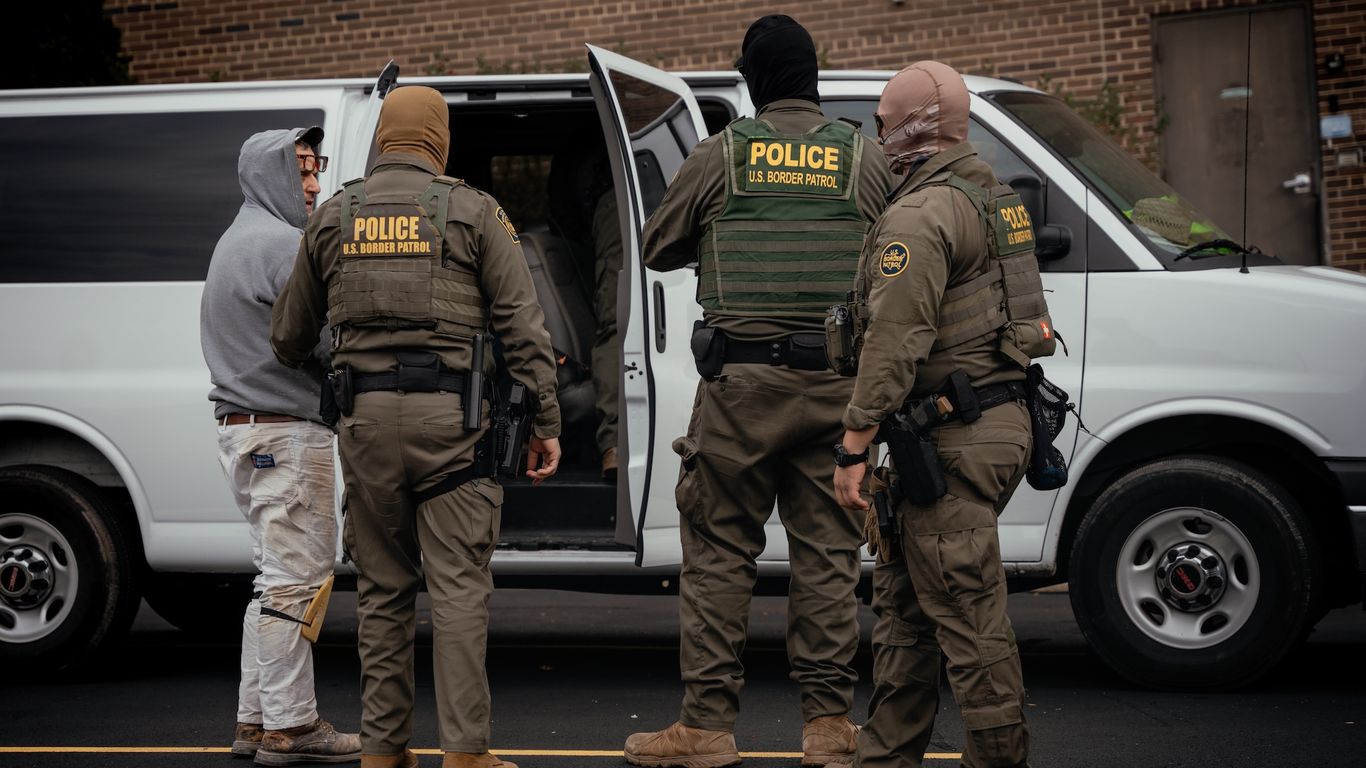 The backs of three federal law enforcement agents next to a man in a gray hoodie and white pants next to a white van with the door open.