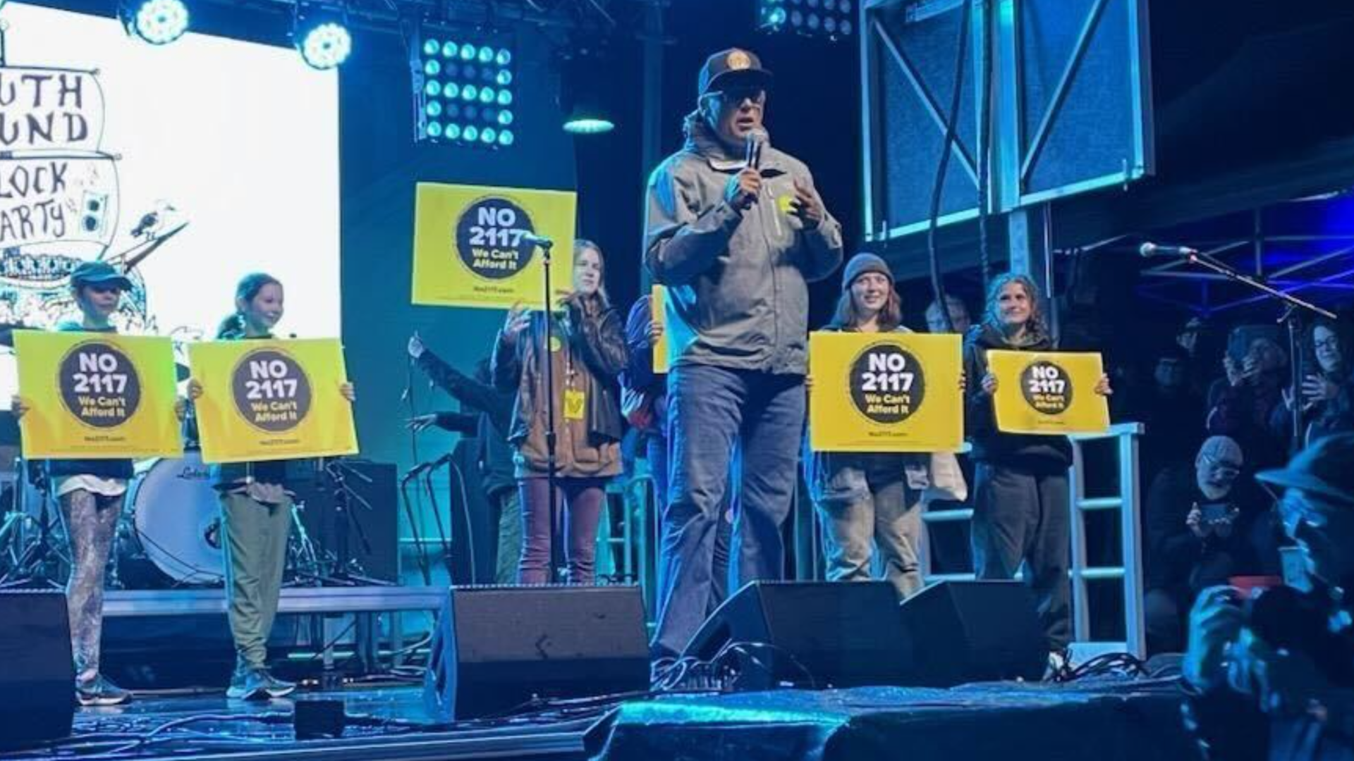 Washington Governor Jay Inslee, in jeans a ballcap and windbreaker, speaks into a microphone on stage surrounded by people holding "No on 2117" signs.