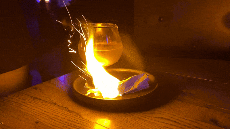 A moving image of a plate full of fire revealing a cocktail on a plate beside a chicken foot.