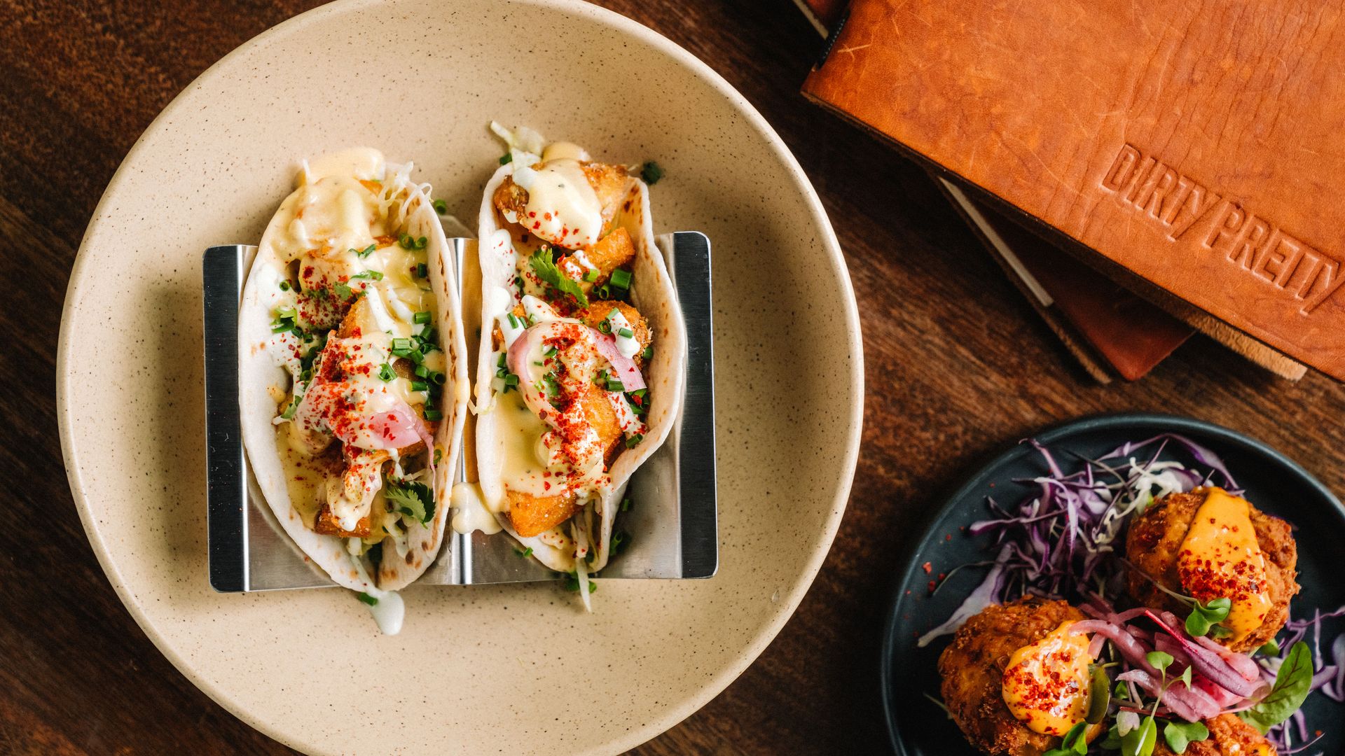 Two loaded tacos with crispy potatoes, creamy sauce, herbs, and radishes sit on a beige plate with a taco holder; nearby, a dark plate holds fried bites topped with orange sauce and greens.