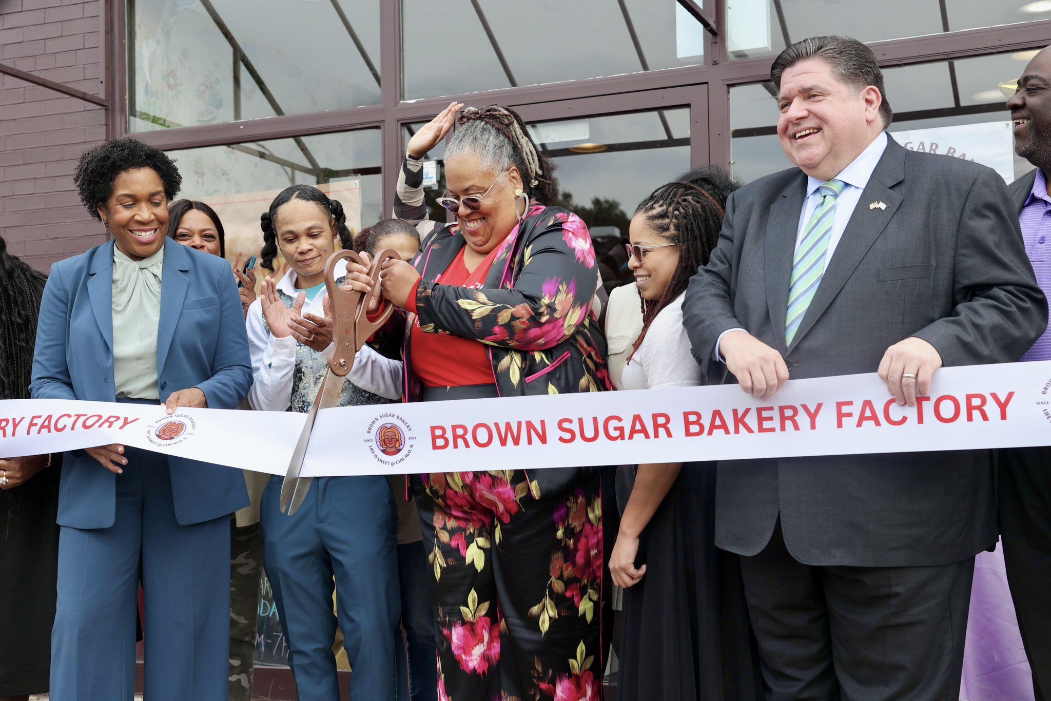 Stephanie Hart stands next to Gov. JB Pritzker with giant scissors, cutting tape that says "Brown Sugar Bakery Factory."