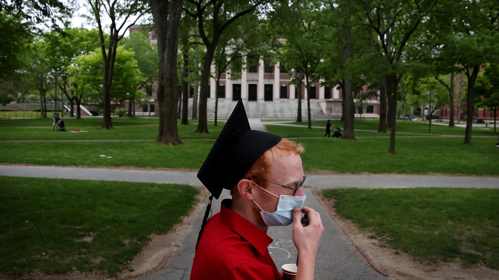 A student in a cap and gown and face mask walks down campus sidwalk