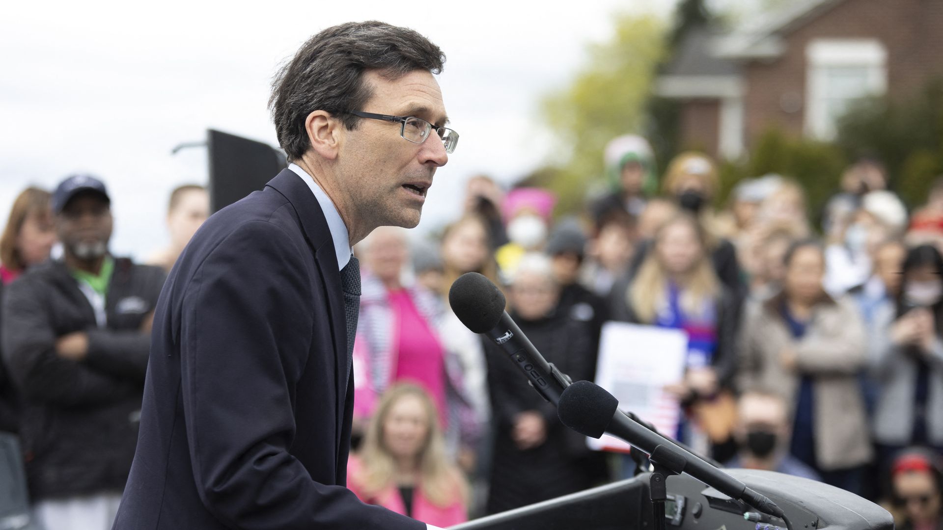 A man with short brown hair and glasses in a dark suit speaks at a podium with a crowd in the background.