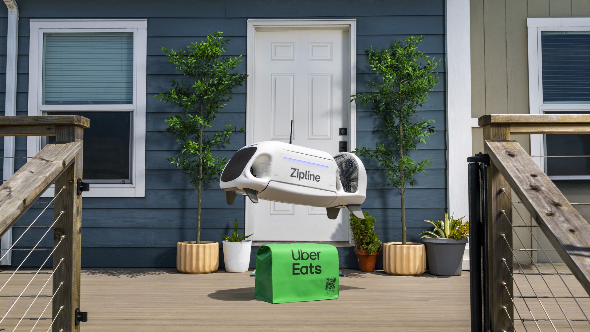 A white Zipline drone delivers a bright green Uber Eags bag to the wooden porch of a blue house with a white door, two potted trees flanking the entrance.