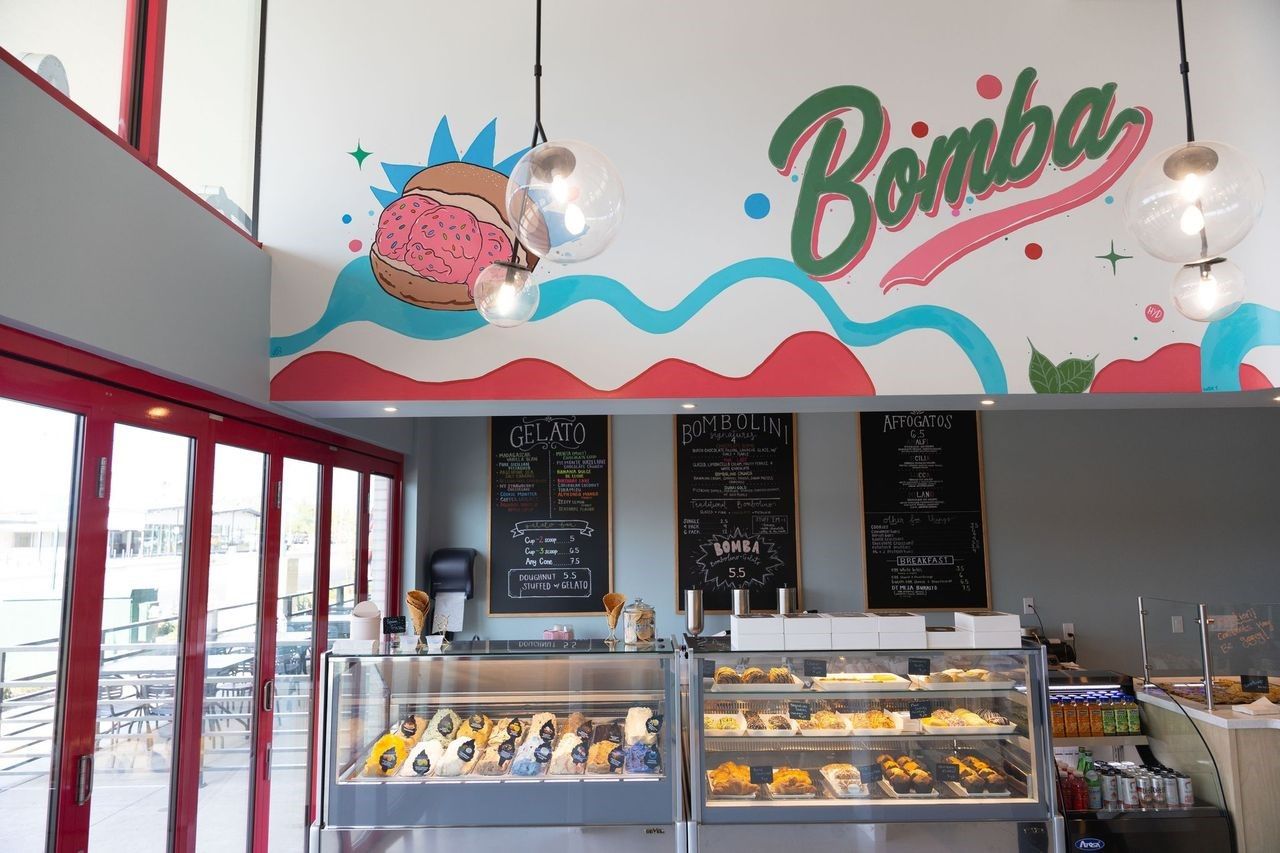 Bright bakery cafe with red-framed windows, a vibrant mural, hanging glass lights, and a pastry display case. Chalkboard menus list Gelato, Bombolini, and Affogatos.