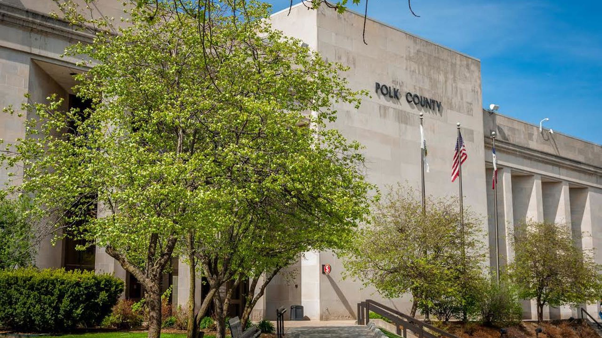 Beige Polk County government building with POLK COUNTY on the facade, green trees in front, a paved walkway, and flagpoles with the U.S. flag and others under a blue sky.