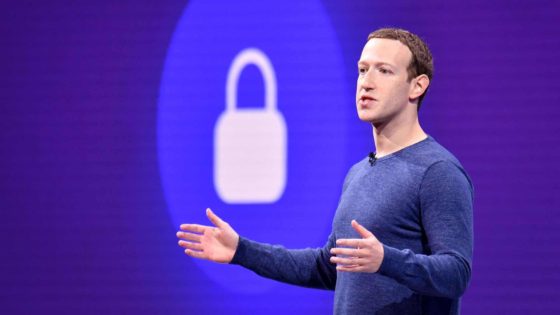 Photo of Facebook CEO Mark Zuckerberg with arms out and a lock icon projected behind him on stage