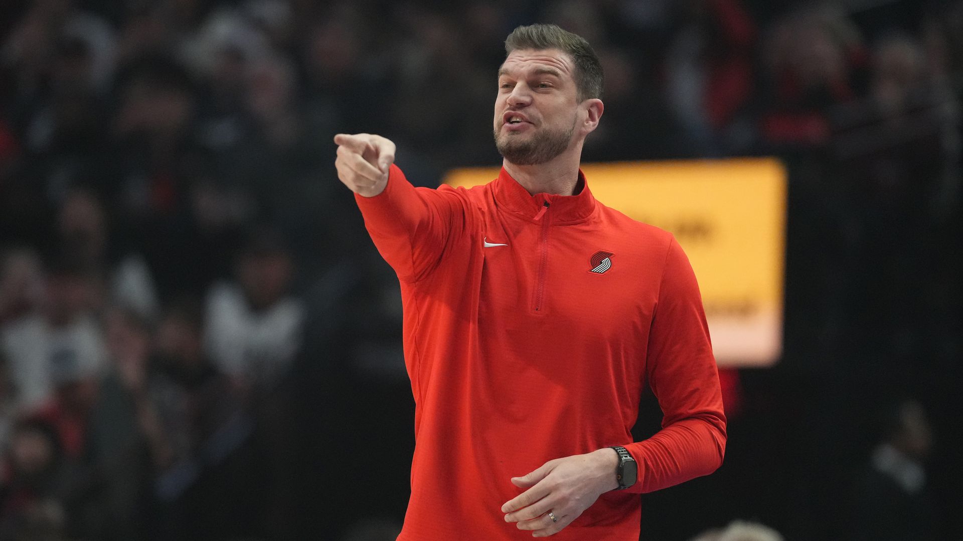Male athlete in a bright red zip-up jacket with a Portland Trail Blazers logo points outward while speaking, in a dim arena with a blurred crowd behind him.
