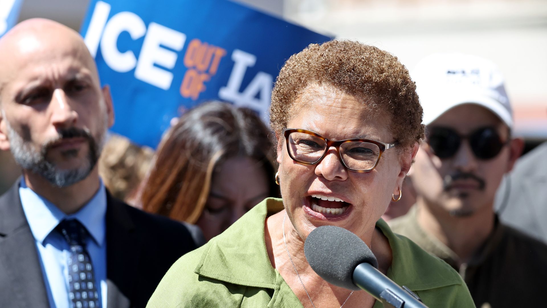 Los Angeles Mayor Karen Bass, wearing tortoiseshell glasses, a green blouse and silver necklace with a diamanté pendant, speaks into a mic as people look on in the background. A blue sign saying in white and orange writing "ICE OUT OF LA" can be seen just behind her. 