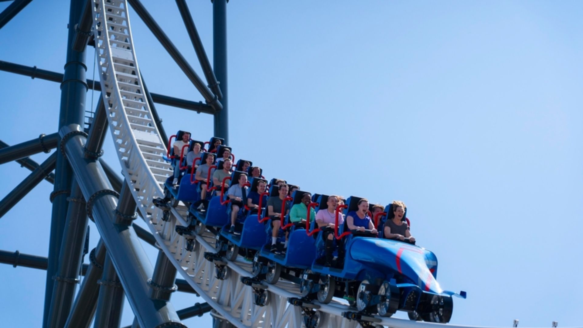 A blue roller coaster train with riders strapped in red restraints travels along a white track beneath a clear blue sky.