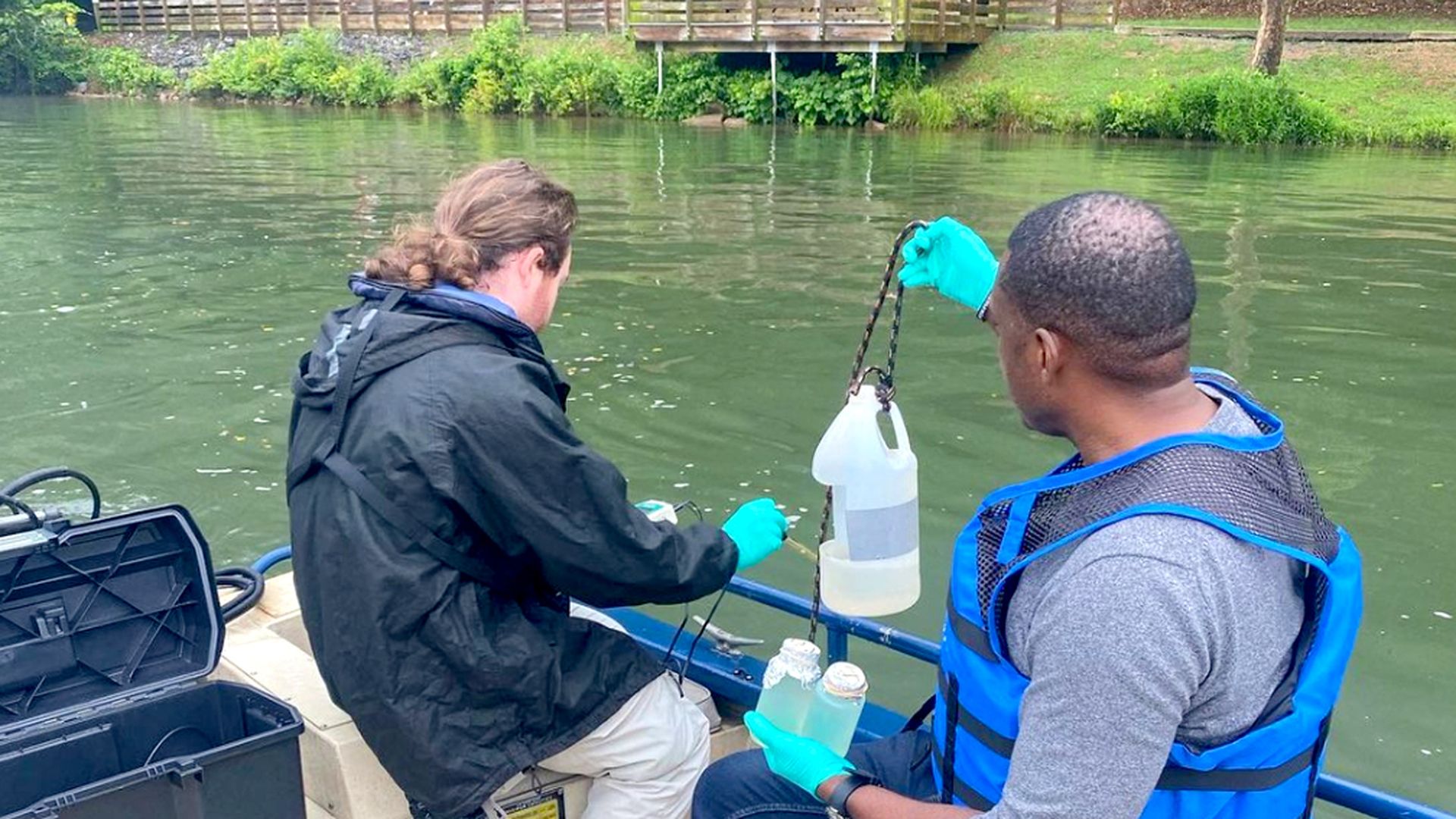 Two men wearing protective gloves in a boat lift jugs containing water samples on the Chattahoochee River