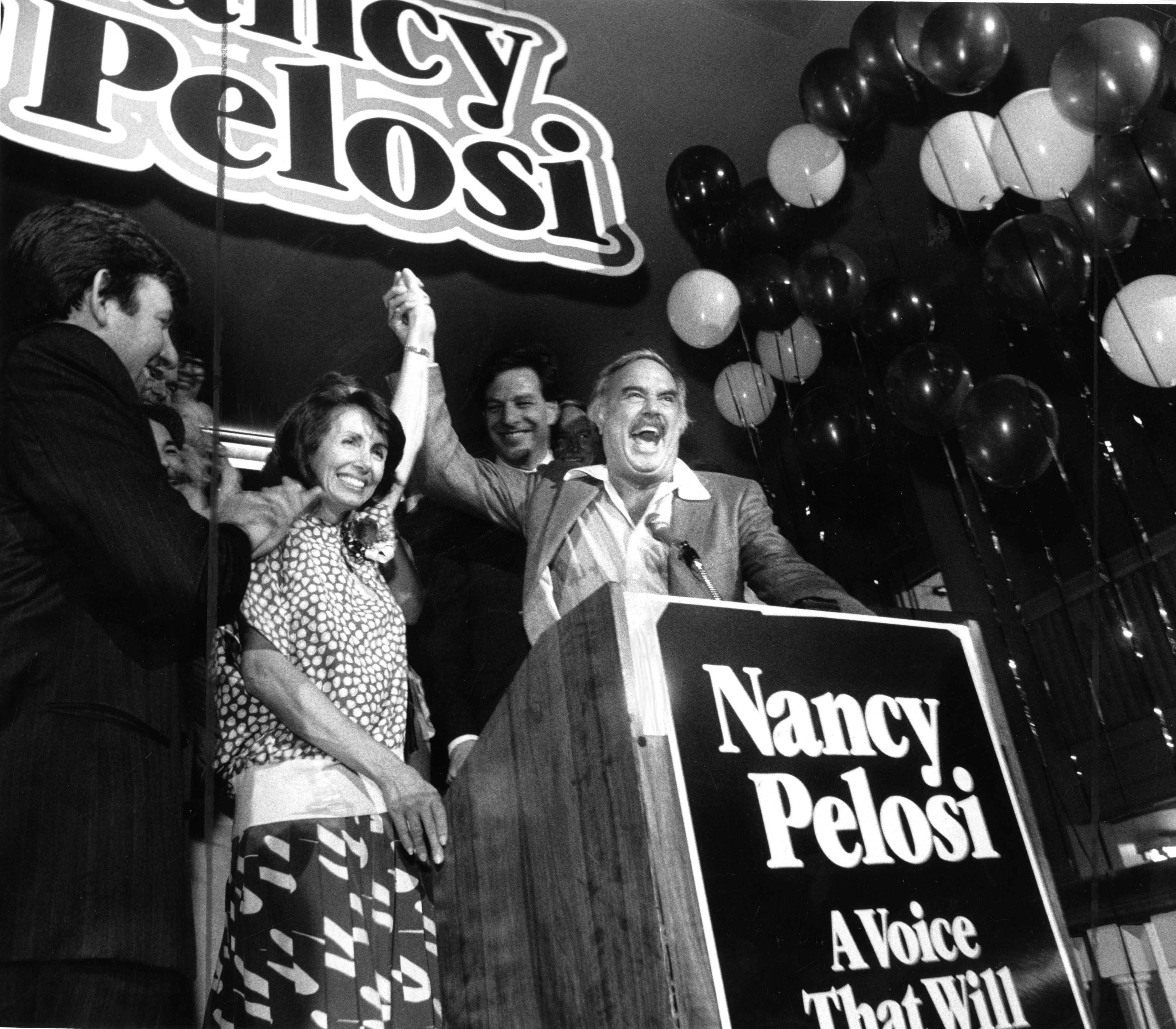 A man holds Nancy Pelosi's hand in the air in this black and white photo as they smile. He stands behind a podium that reads "Nancy Pelosi" as balloons float nearby.