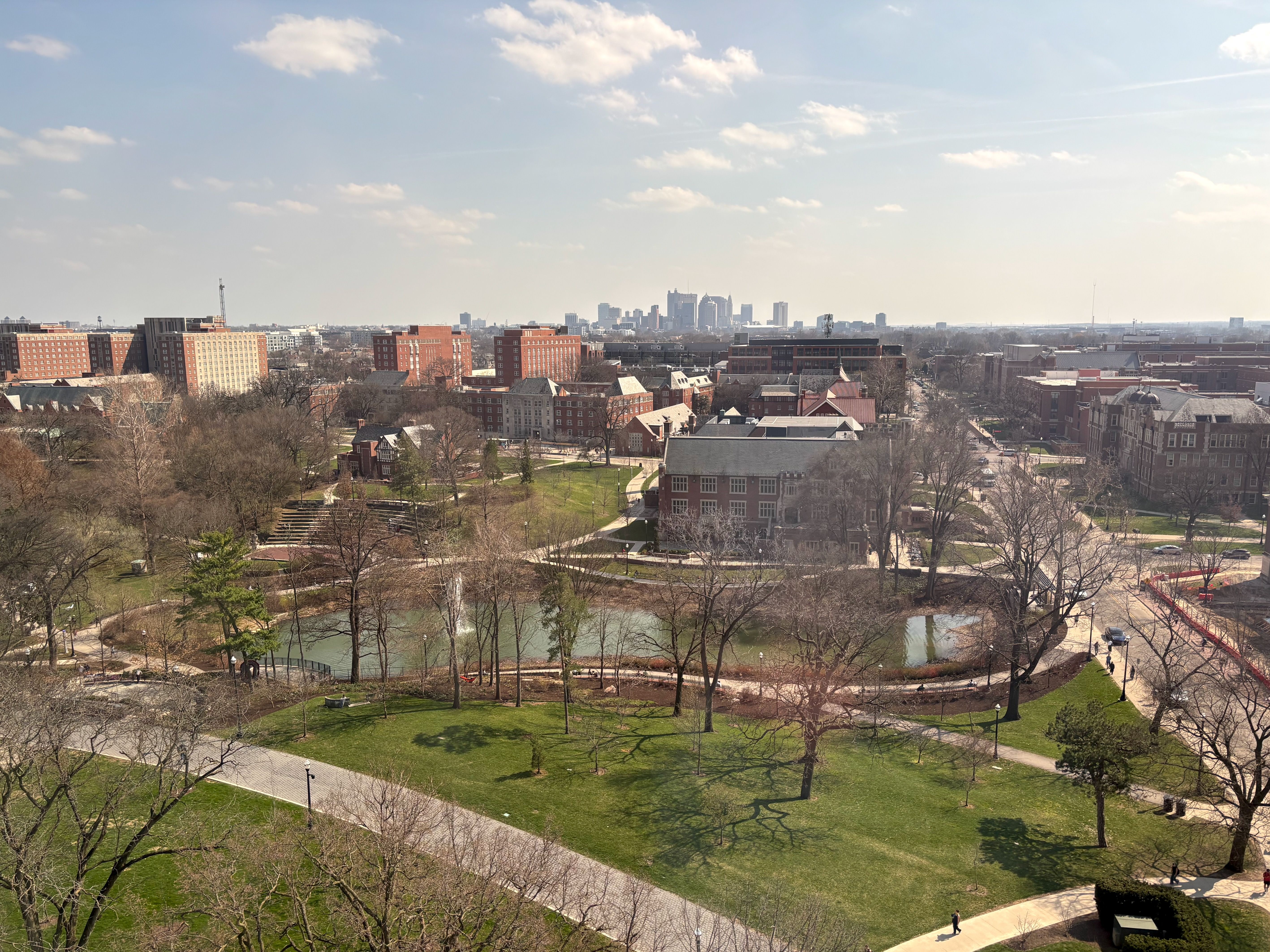 Panoramic view of Ohio State University's south campus on a sunny day: green lawns, bare trees, winding paths, and a pond with a fountain. Red-brick buildings line the mid-ground; distant skyline with tall towers.