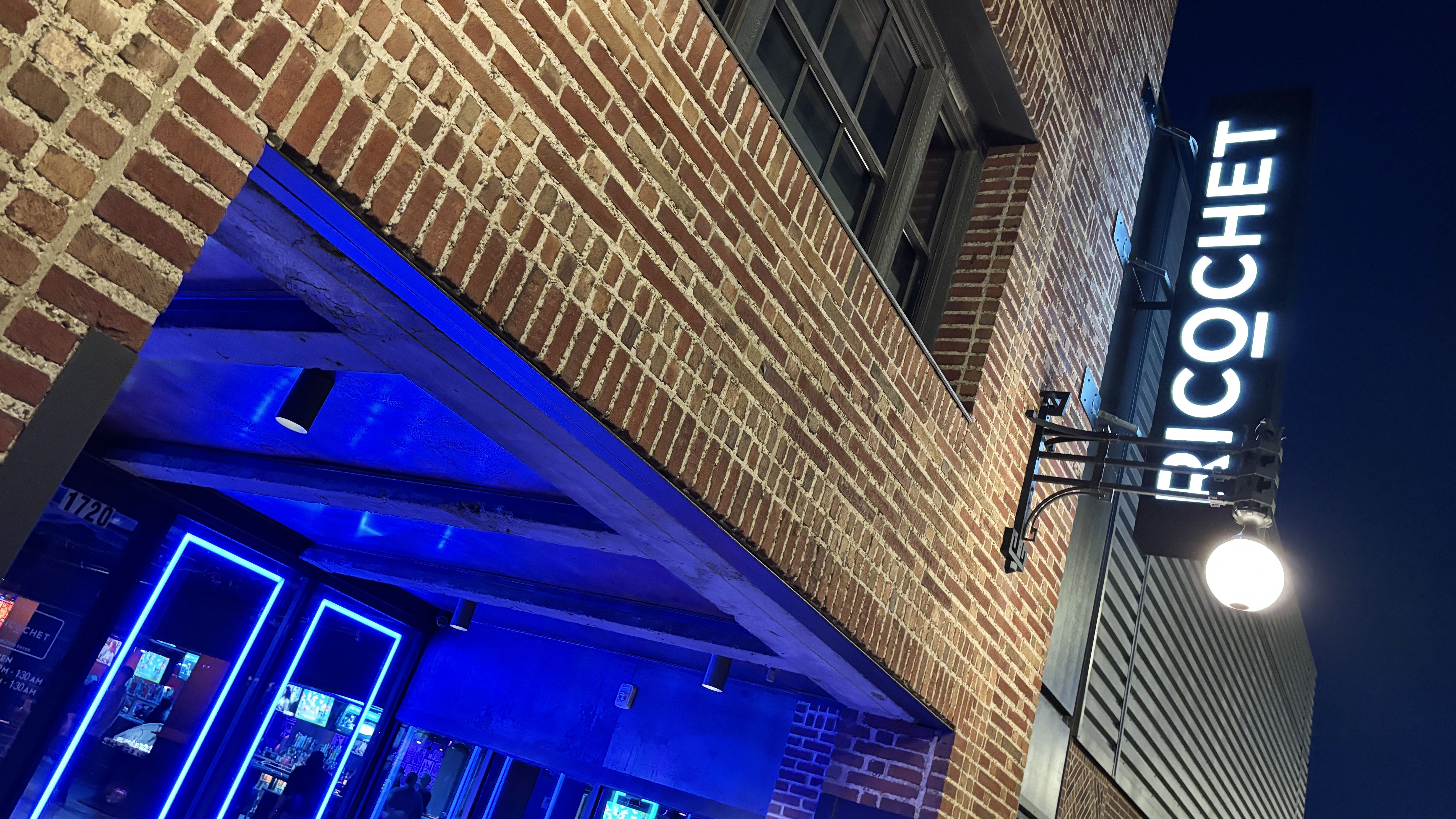 Night view of brick building with blue neon-lit entrance and a bright white sign reading "RICOCHET" against a dark sky.
