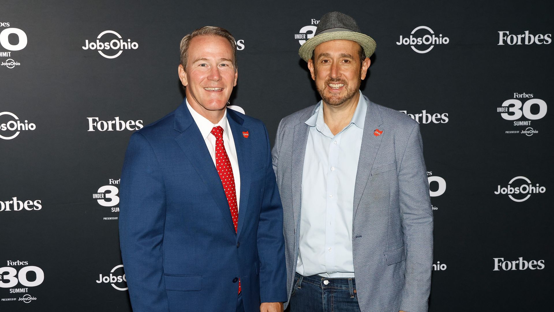 Man in blue suit next to man in jeans, gray jacket and fedora, standing in front of black wall with Forbes and JobsOhio logos.