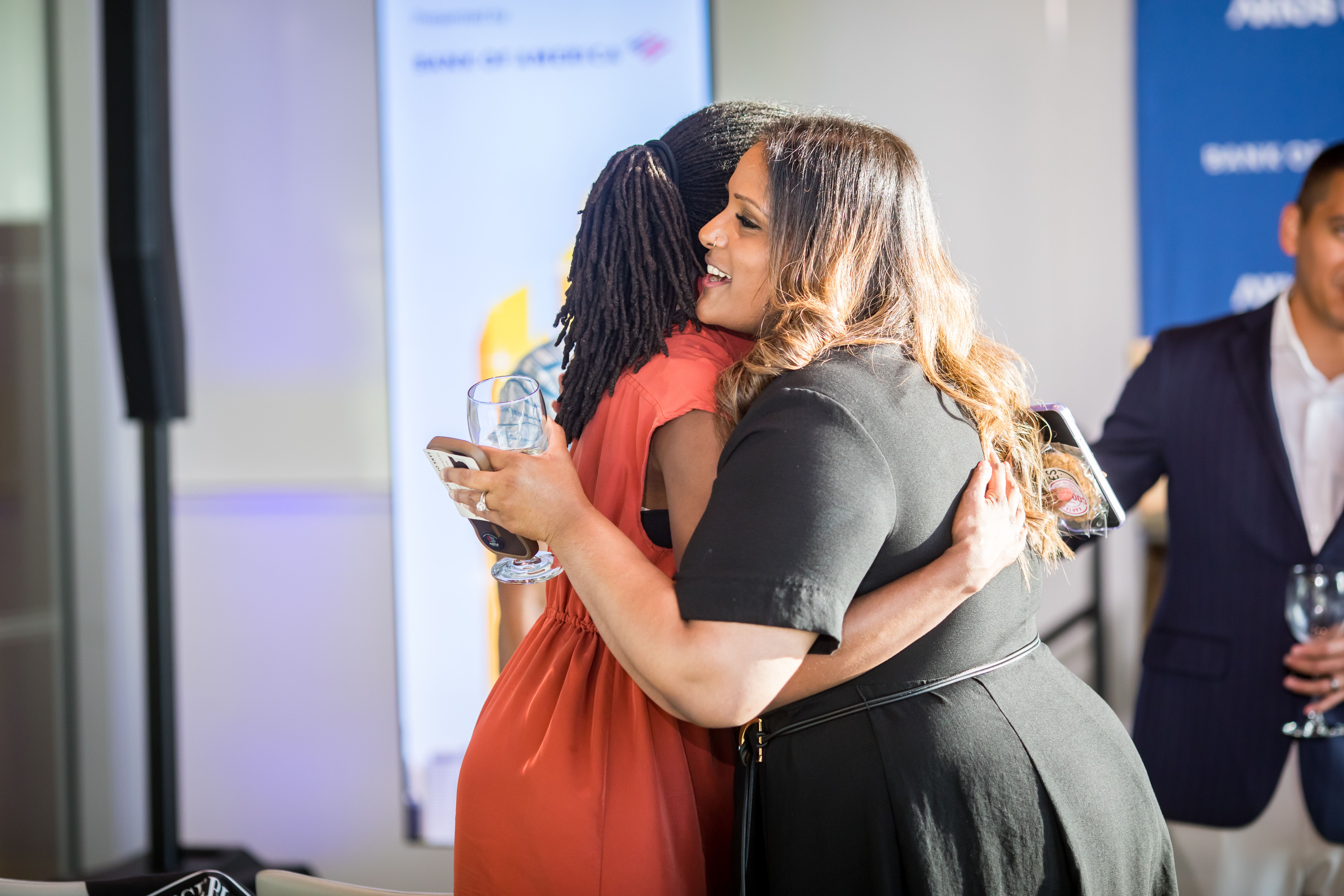 Two women hug at an event; one in an orange dress holds a wine glass, the other in black. A man in a suit stands nearby, with blue banners in the background.