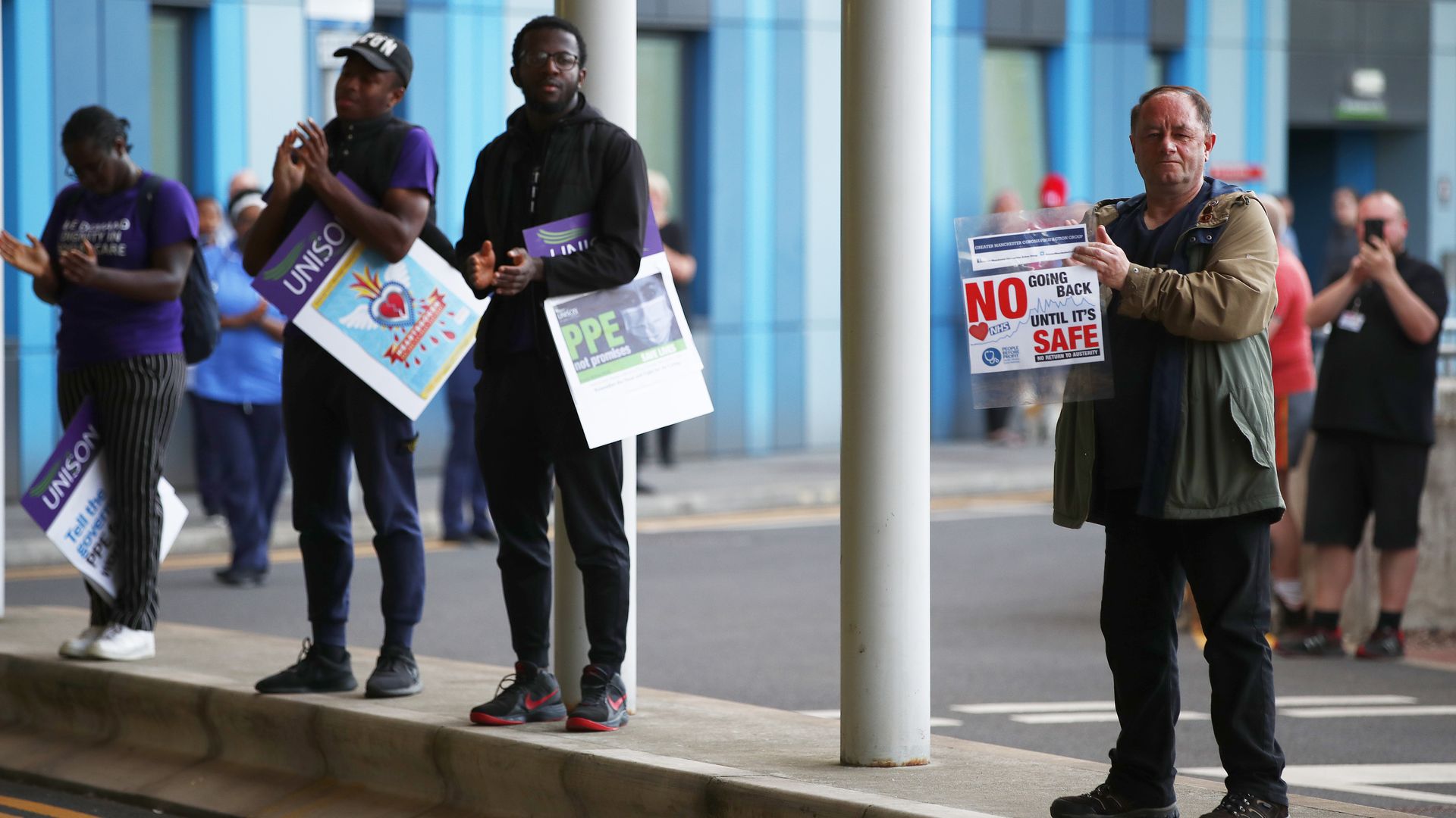 In this image, people stand in a row outside and clap, some holding signs