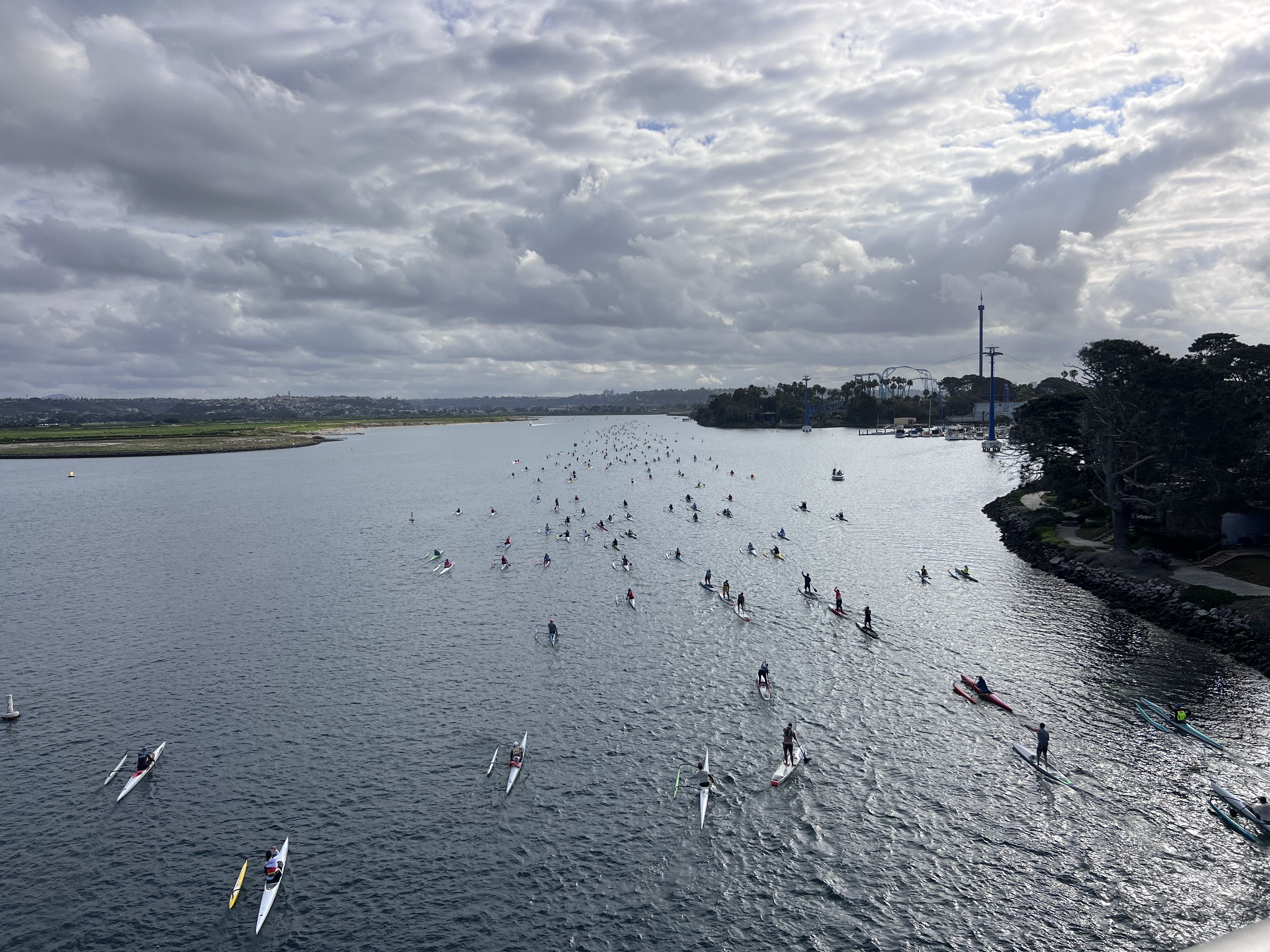 Wide river scene under cloudy sky with hundreds of people kayaking and paddleboarding along the water, bordered by green land on the left and trees on the right.