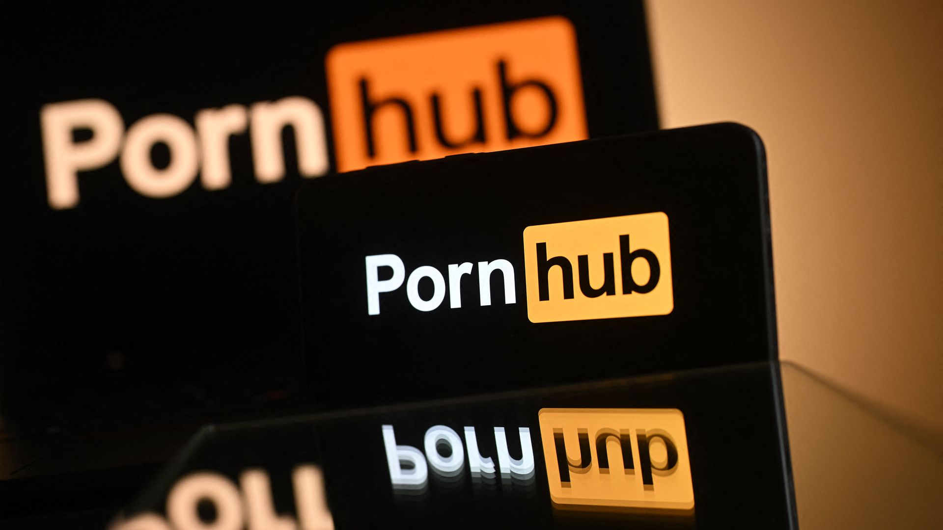 Close-up of black screens showing the word "Pornhub" with orange badge icons; warm lighting, out-of-focus foreground, bold white text on dark background.
