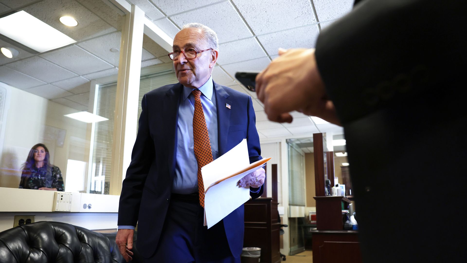 Senate Majority Leader Chuck Schumer, wearing a blue suit, light blue shirt, rust tie and glasses, holding sheets of paper as he walks to a press conference.