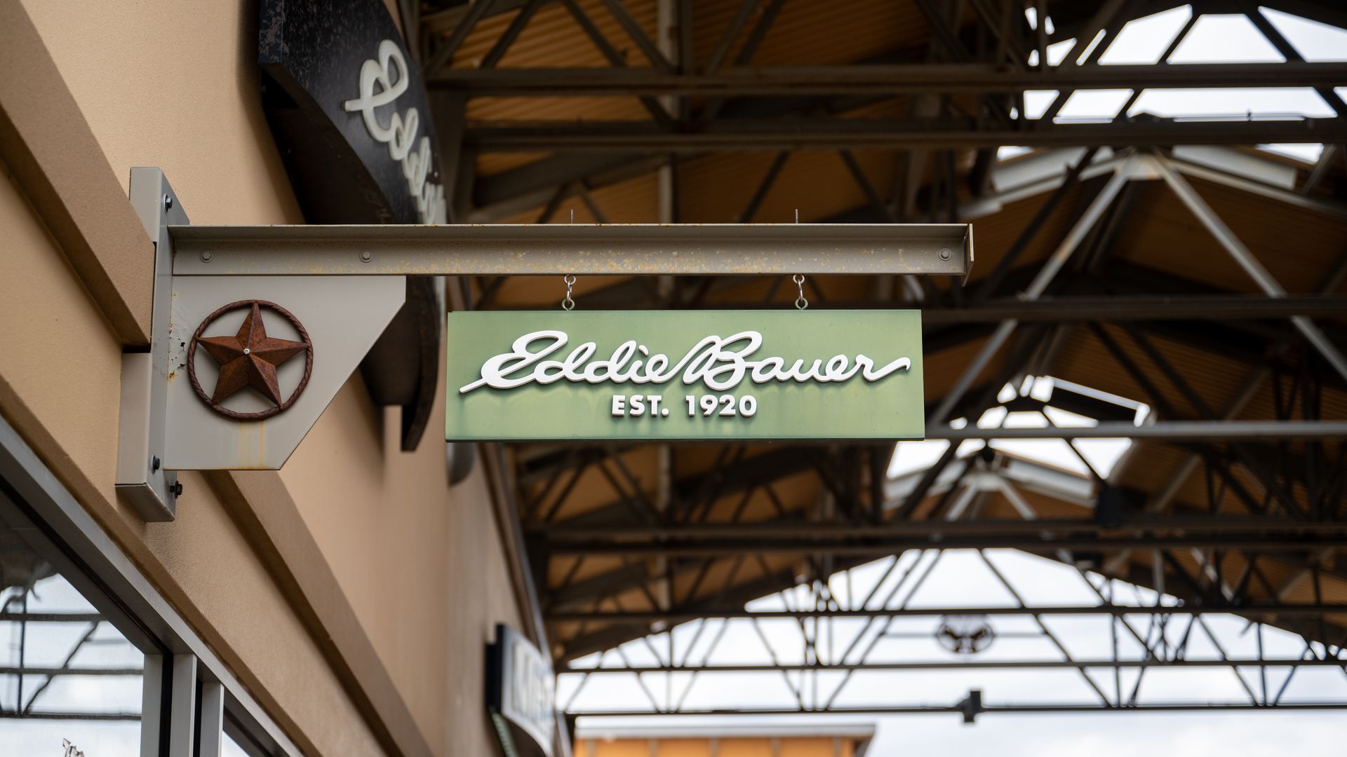 Green rectangular sign with white cursive text "Eddie Bauer EST. 1920" hanging under a beige building awning with exposed metal roof beams and a rusted star decoration on the mounting bracket.