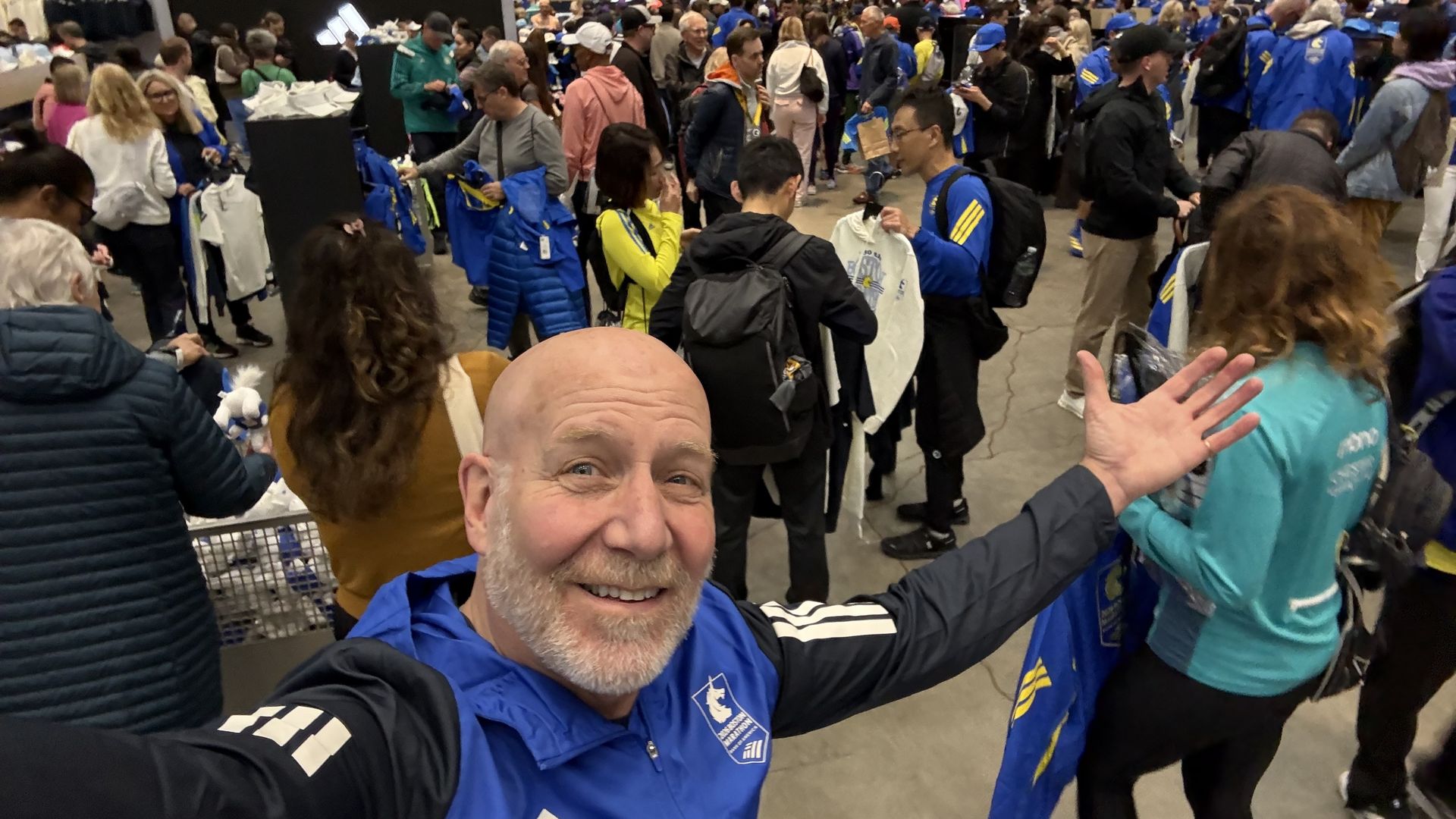 Busy indoor expo with many attendees shopping in athletic gear. A smiling bald man in blue jacket takes a selfie in the foreground; crowd in blue and yellow outfits fills the background.