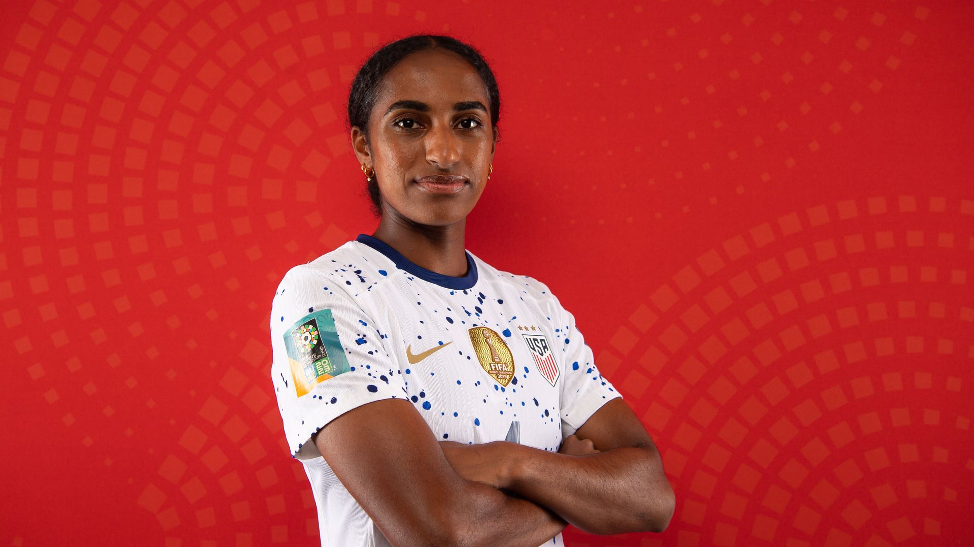 A player on the U.S. women's national soccer team wearing a white jersey with a splattered blue design stands with her arms crossed and a slight smile. 