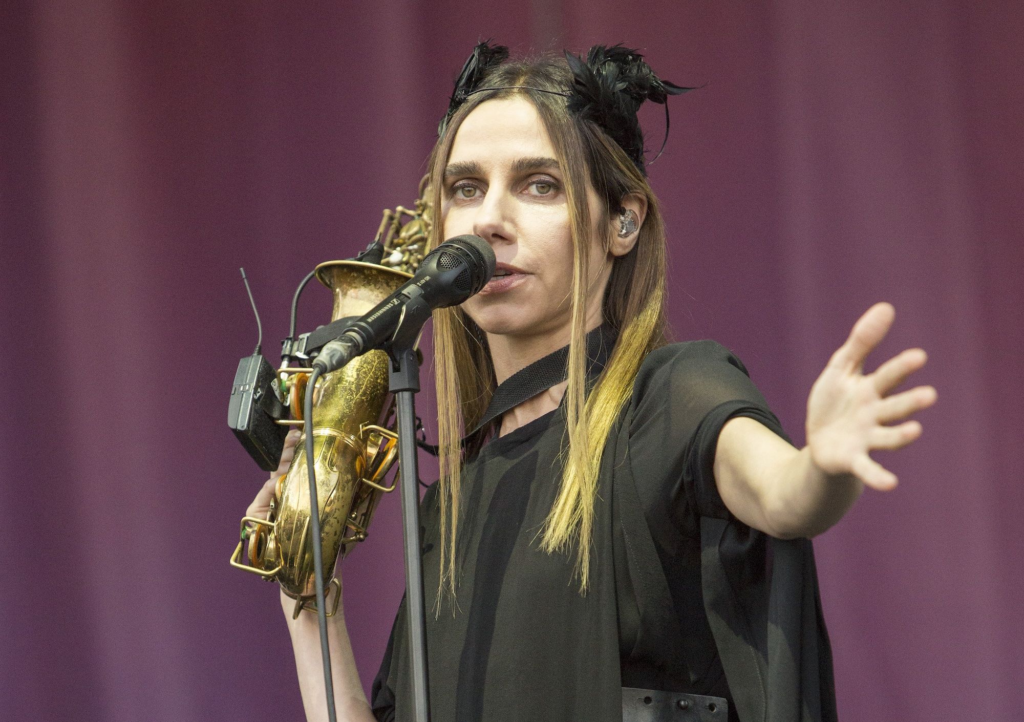 PJ Harvey in black dress with black bows in her hair holding a brass instrument singing into microphone.