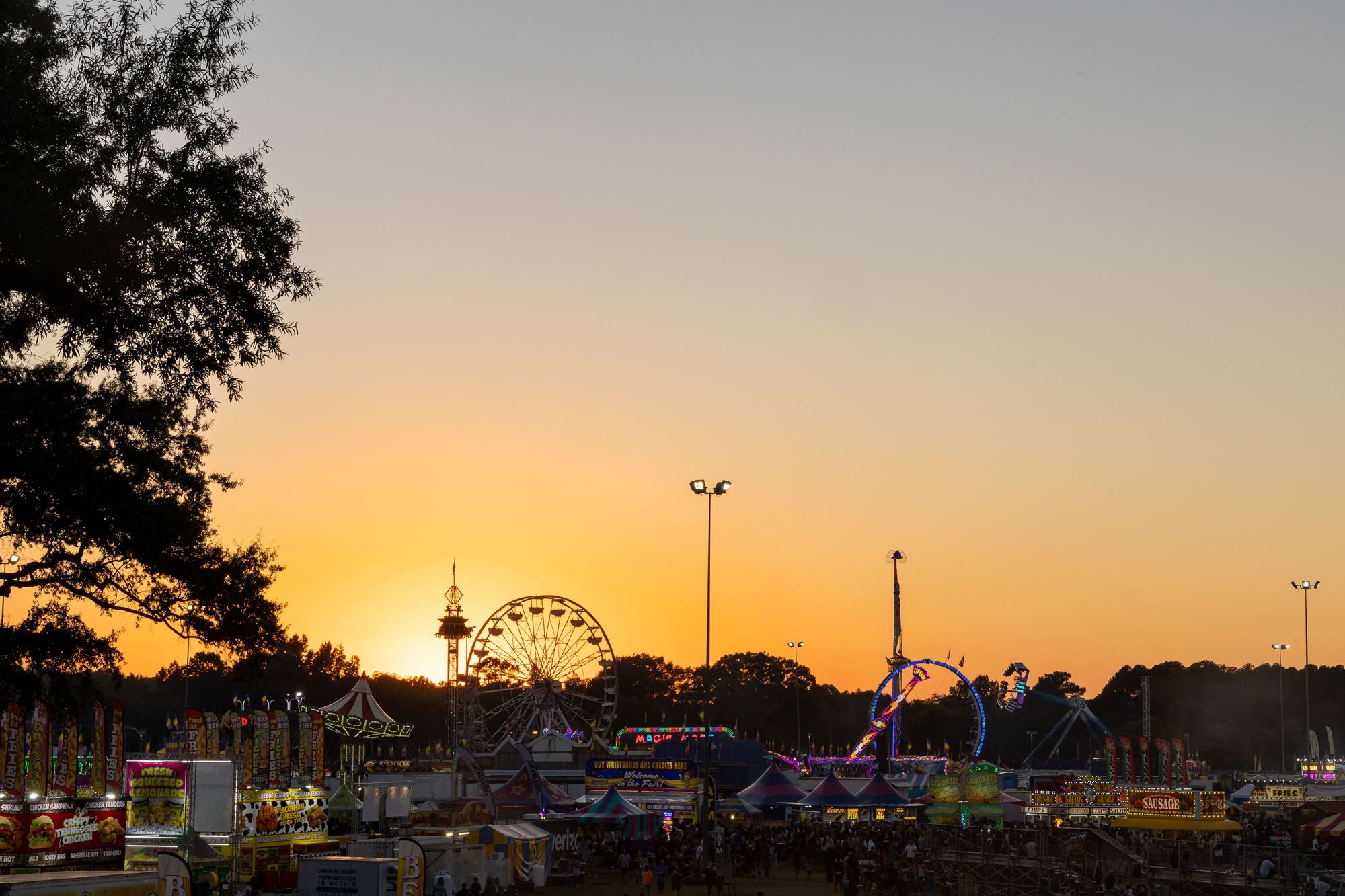 Carnival at sunset with dark tree silhouettes on the left, a large ferris wheel and colorful stalls along the foreground, warm orange sky fading to pale blue.