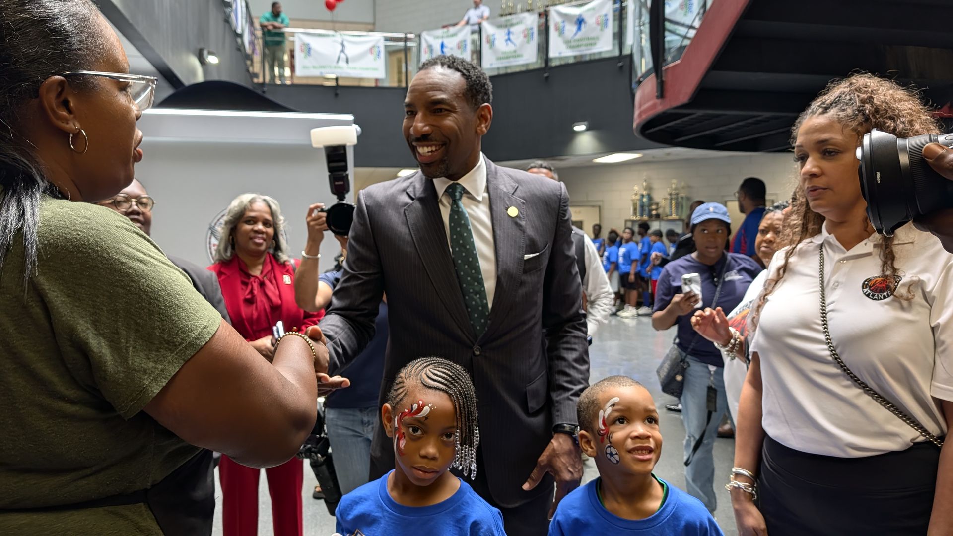 Smiling man in a gray suit and green tie shakes hands with a person in a green top at a busy indoor event. Children in blue shirts with painted faces stand nearby as photographers observe.