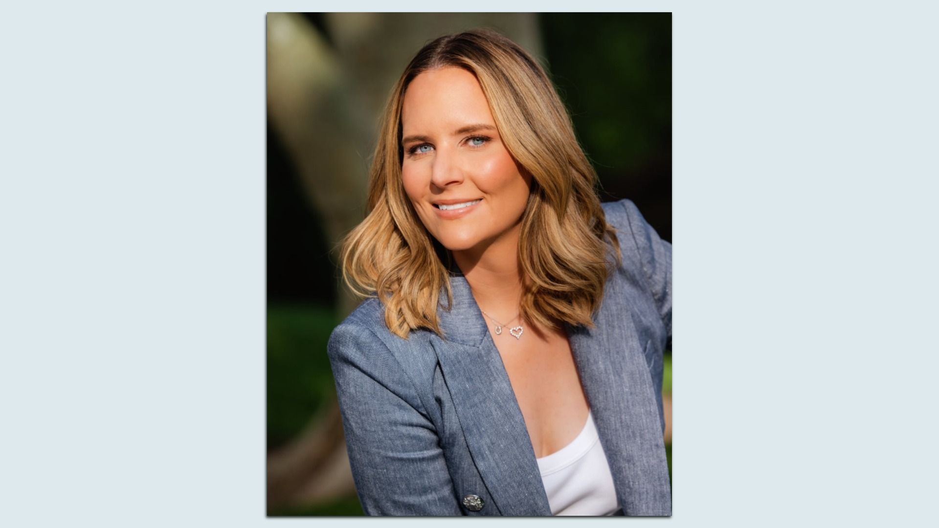 Smiling woman with shoulder-length blonde hair, blue eyes, wearing a gray blazer and white top, outdoors with blurred green and dark background.