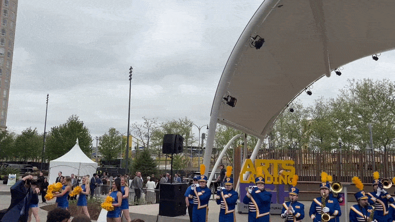 A marching band playing in front of a large canopy and stage with yellow bridges in the background