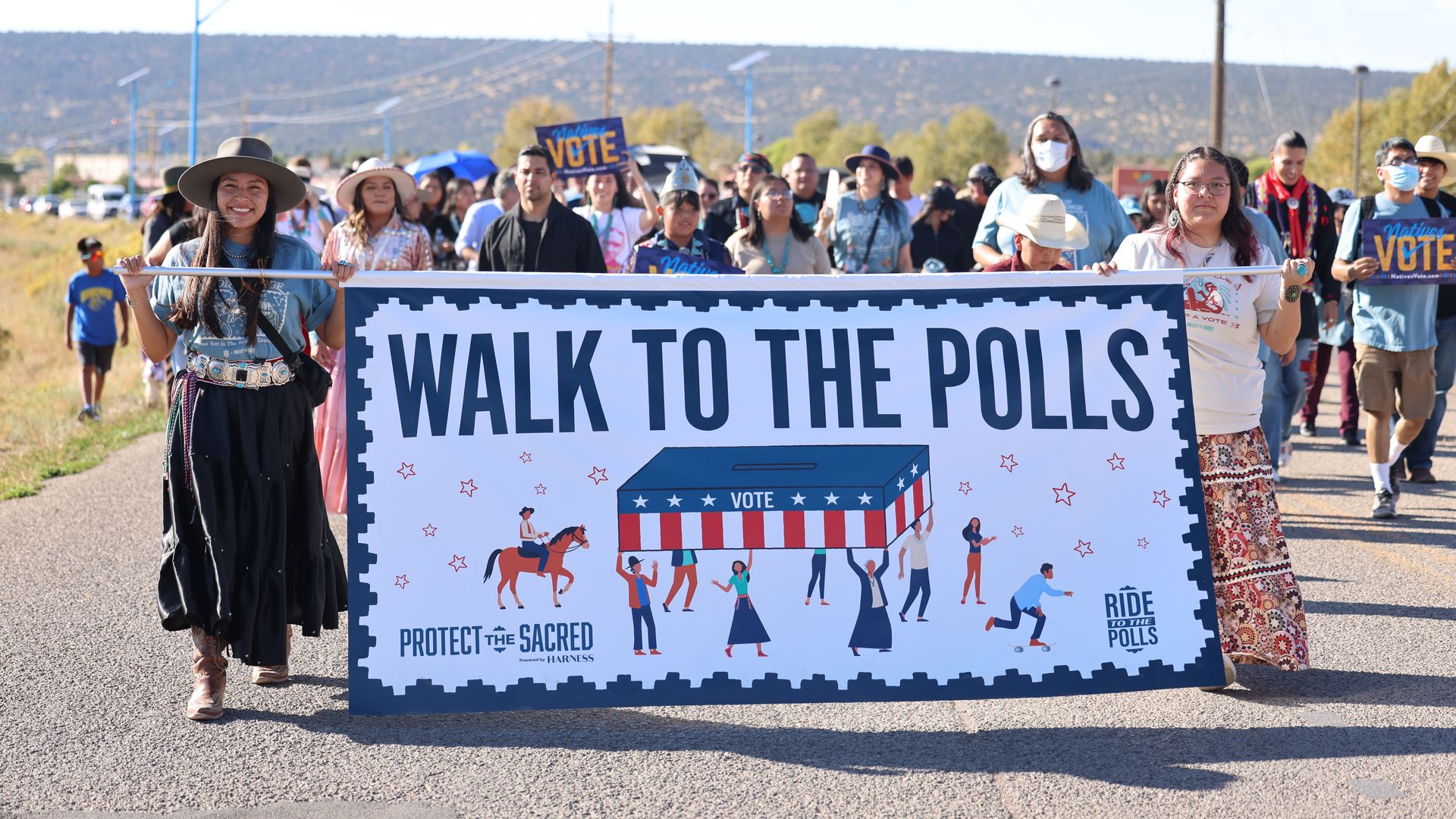 Group of people walking outdoors on a sunny day holding a large banner that reads "Walk to the Polls" with illustrations and messages supporting voting and protecting the sacred.