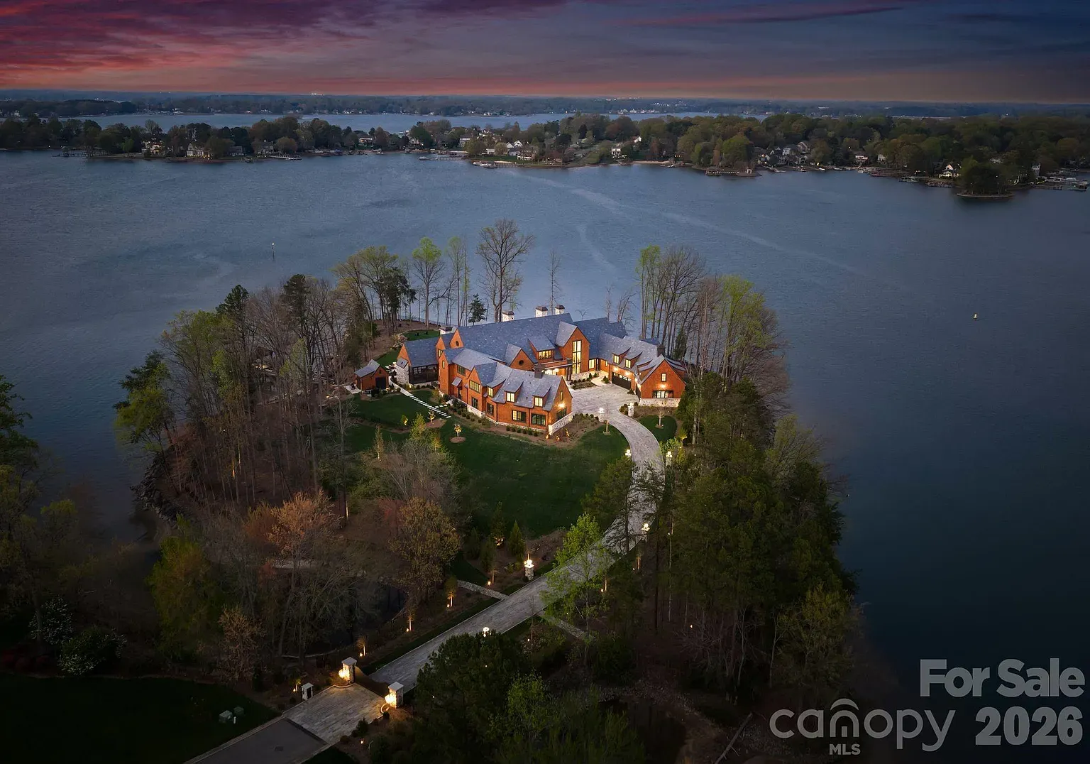 A large brick mansion sits on a wooded private island in a calm lake, linked by a curved driveway. Twilight sunset paints the sky orange and purple, with distant shorelines visible.