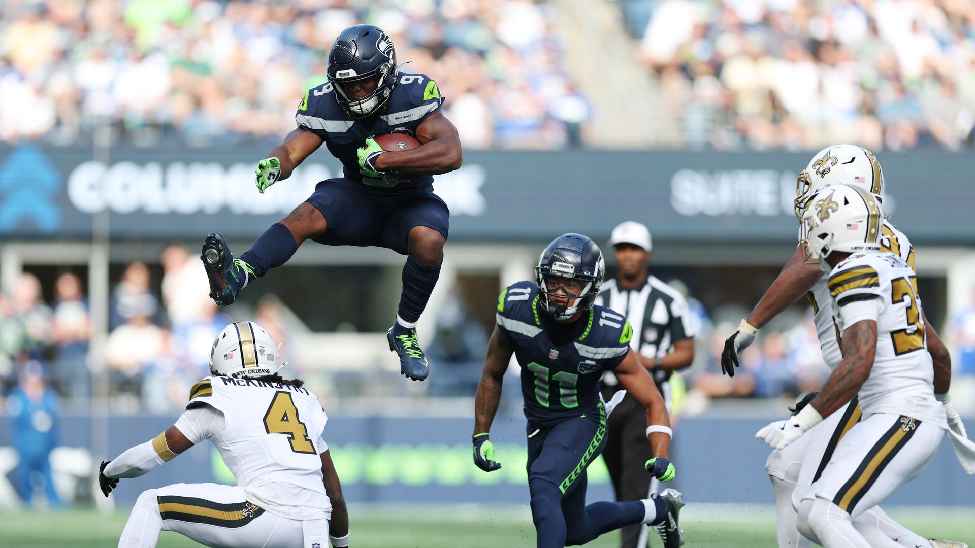 A football player in dark blue leaps over a player in white. 