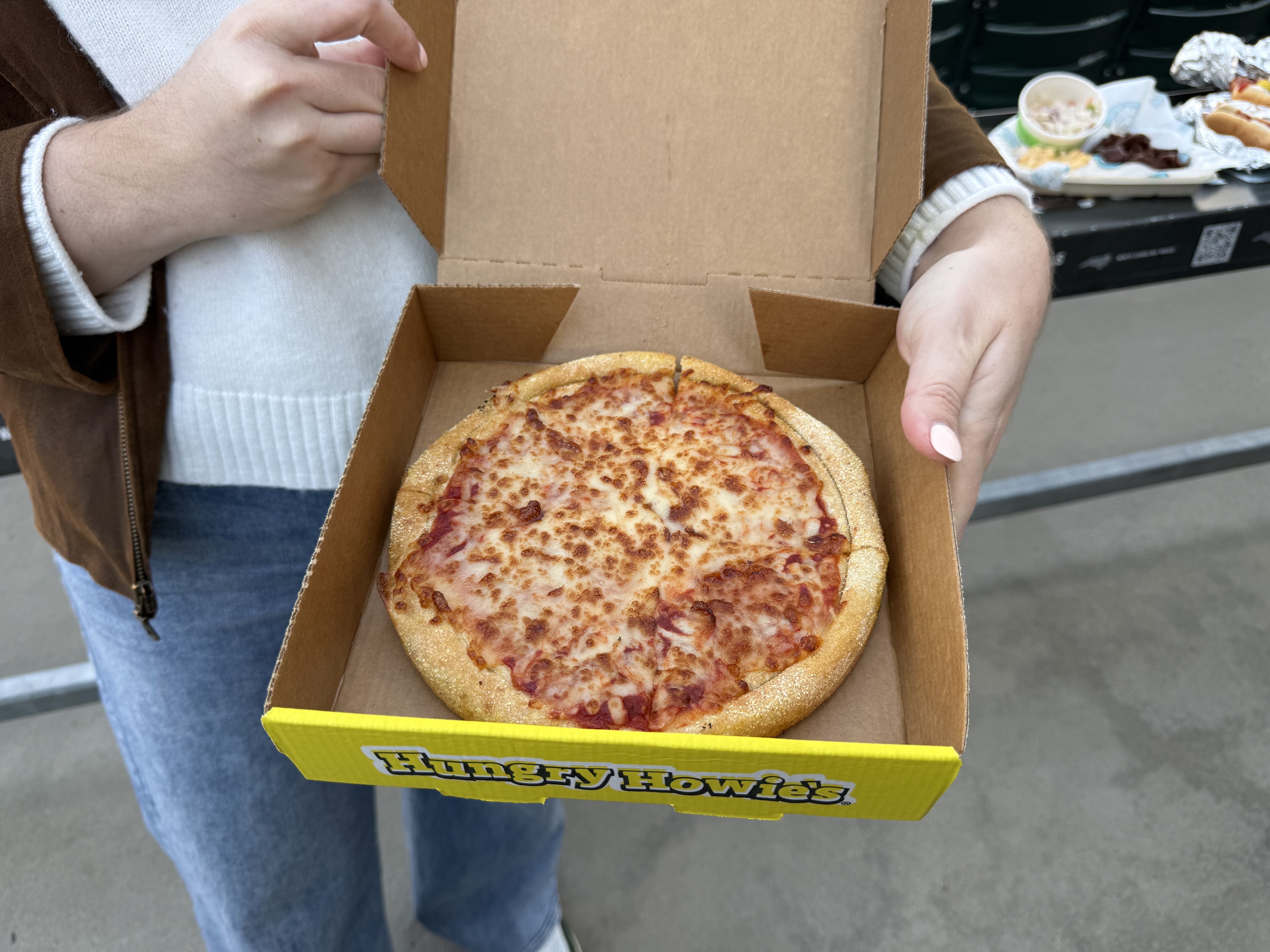 Person in a brown jacket and white sweater holds an open Hungry Howie's pizza box, displaying a pepperoni and cheese pizza with a thick golden crust outdoors.