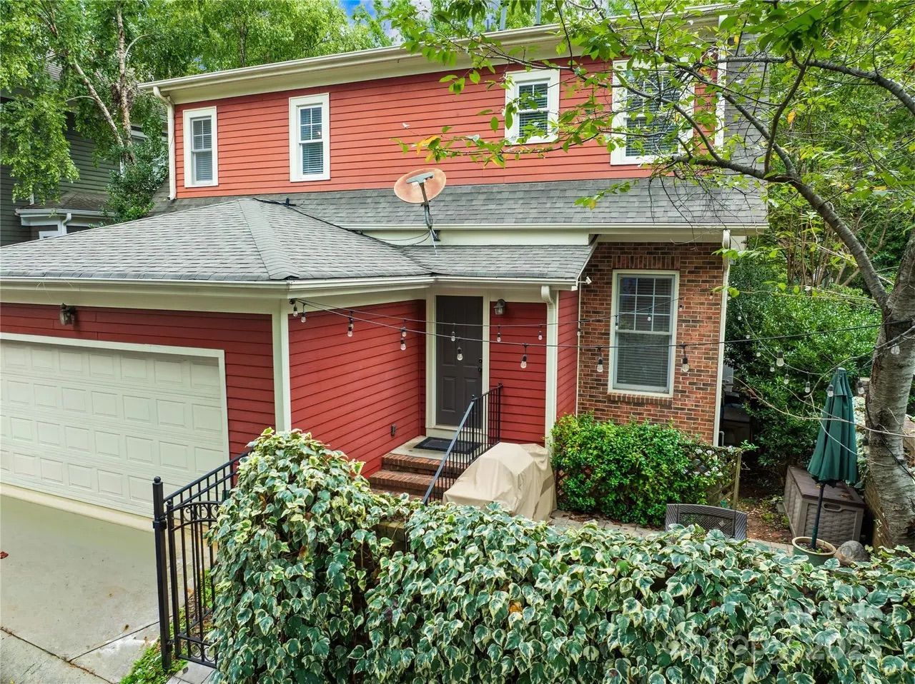 Red two-story house with white trim, gray roof, white garage door, black front door, surrounded by green bushes and trees, with string lights and covered outdoor grill.