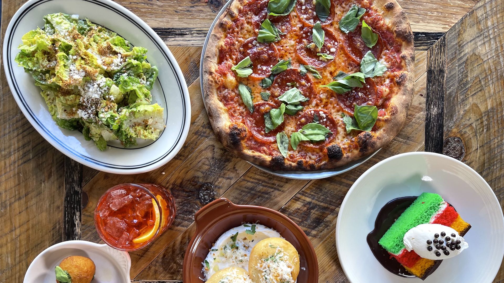 Table with a pepperoni pizza topped with basil, Caesar salad, three fried arancini, a drink with ice and lemon, fried stuffed balls with sauce, and a colorful layered dessert with whipped cream.