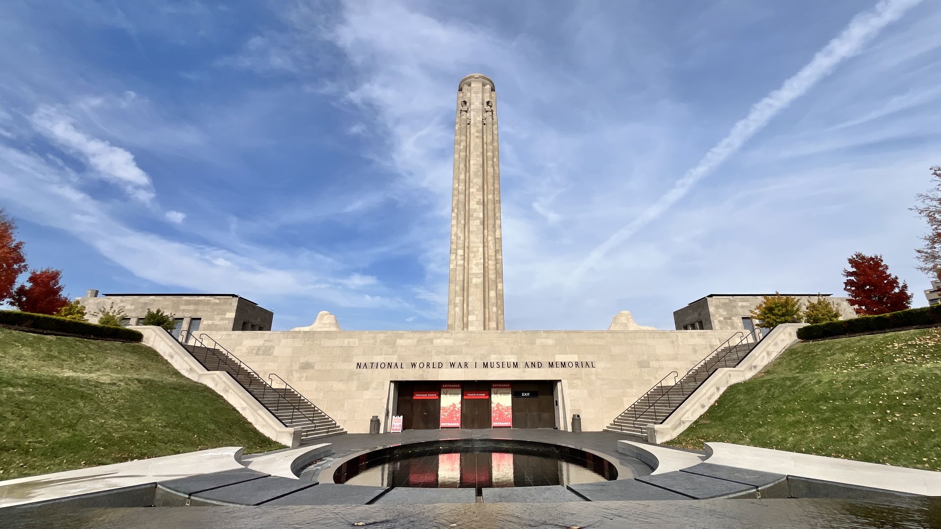 Wide shot of the National World War I Museum and Memorial with a tall central tower, beige stone facade, symmetrical staircases, green lawns, and a circular water feature under a blue sky.