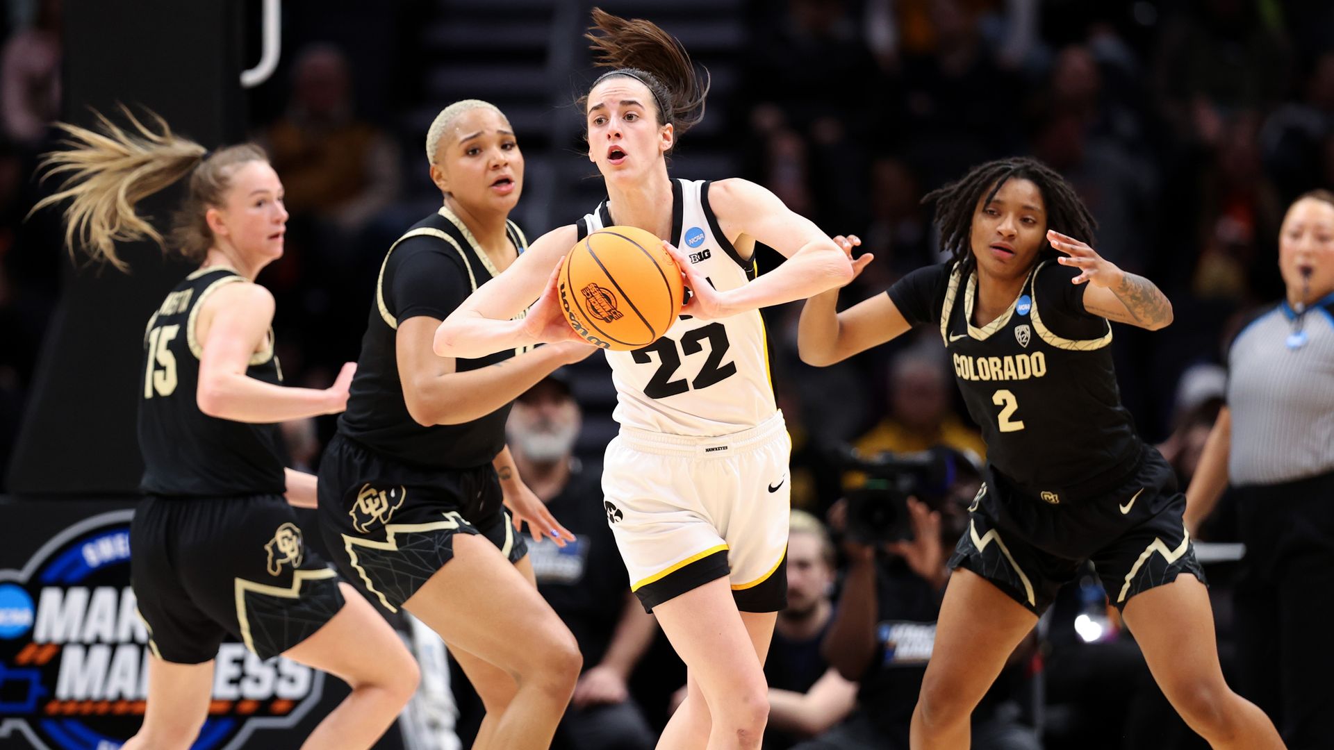 Four women basketball players during a college game, with one woman in a white jersey holding the ball in the foreground and three players in black jerseys in the background look on. 