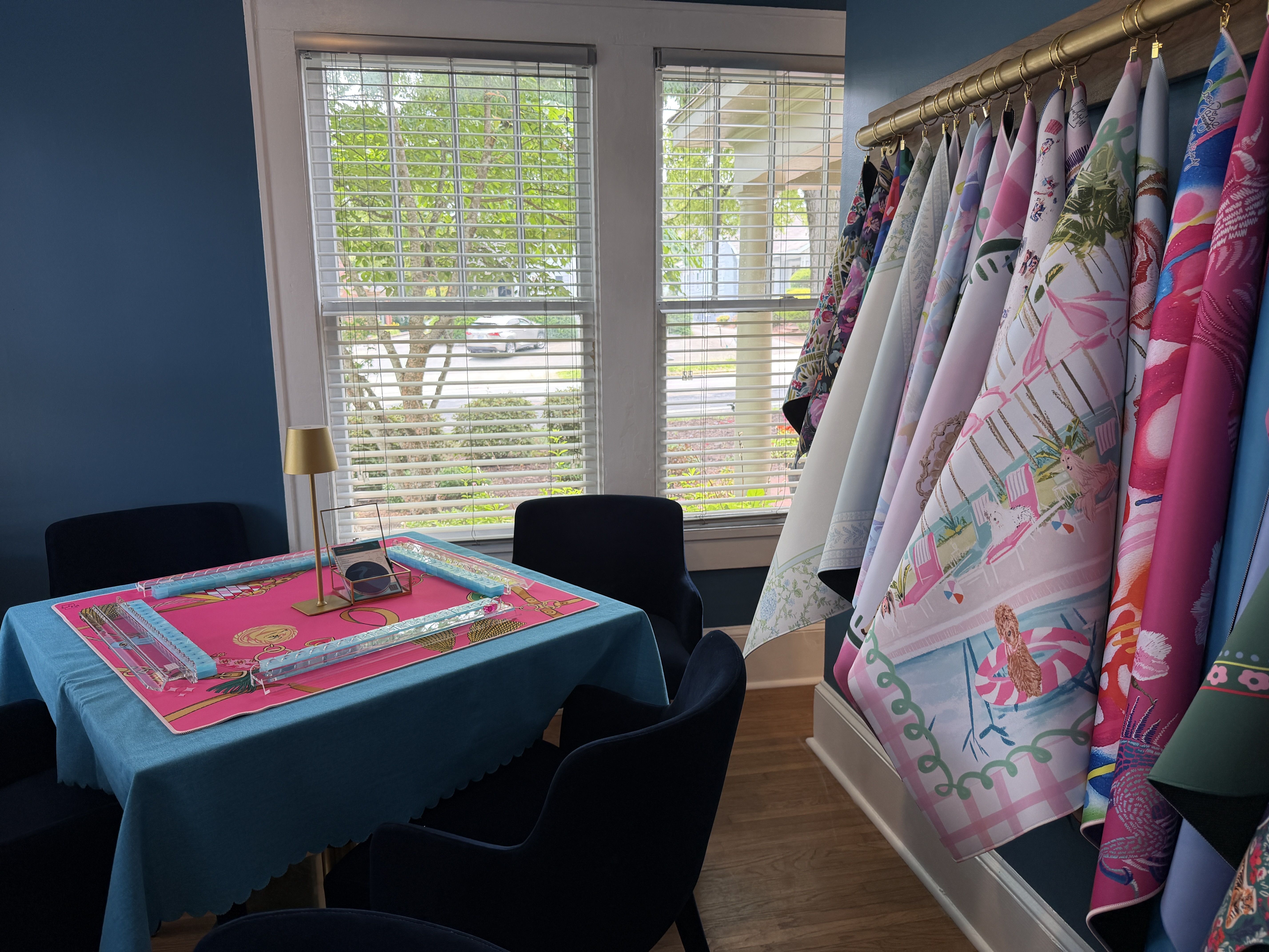 Studio craft room with blue walls and a large window with white blinds. A turquoise table with a bright pink mat sits at left, while colorful tropical fabric bolts hang on a rod along the right.