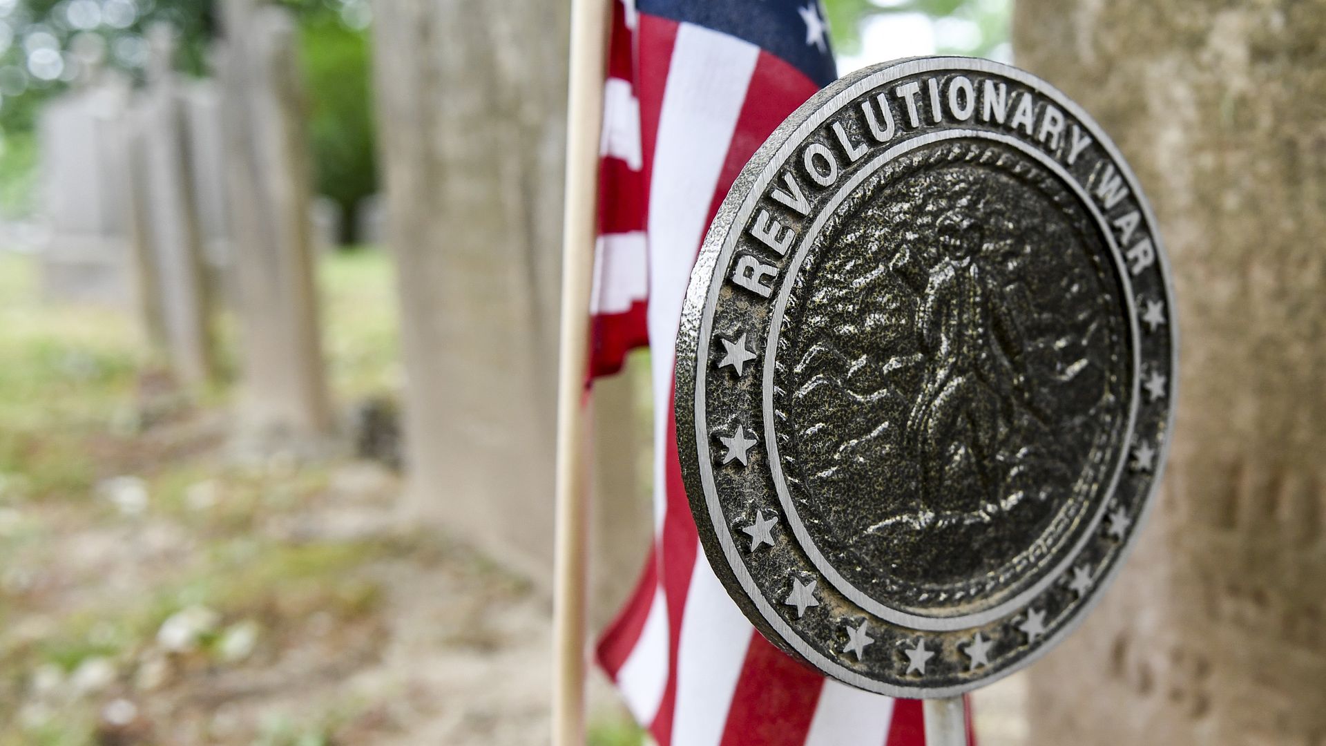 Close-up of a silver commemorative grave marker with the inscription "Revolutionary War" around its rim, worn and weathered, set against an American flag; blurred gravestones and trees in a cemetery background.